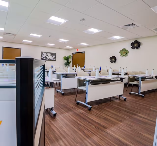 A well-lit room with multiple tables and chairs arranged in rows, each table set with a white napkin and a blue water bottle. The room has wood flooring, white walls decorated with flower-shaped wall art, and a ceiling with recessed lighting. There is a door at the back and a potted plant near it.