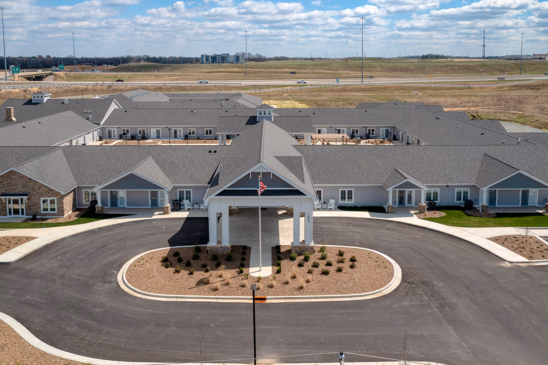 Aerial view of Cedarhurst Senior Living of Owensboro showing a large, single-story building with a covered entrance and an American flag in front. The building has multiple sections with gray roofs and light-colored walls, surrounded by paved driveways and landscaped areas. In the background, there is a highway and open fields under a partly cloudy sky.