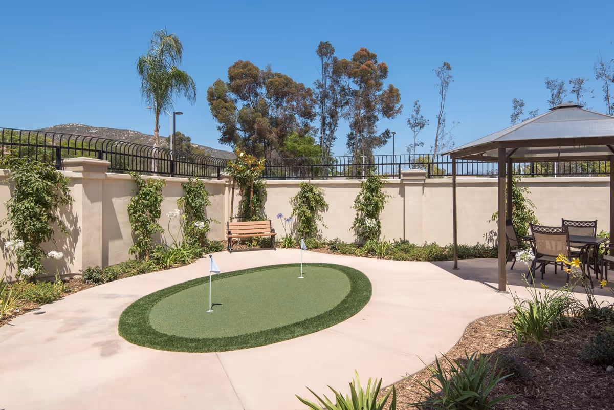Outdoor courtyard area with a small putting green featuring two holes with flags, surrounded by a concrete pathway. There is a wooden bench against a beige wall with climbing plants and greenery. To the right, there is a shaded gazebo with a table and chairs. Trees and a clear blue sky are visible in the background.