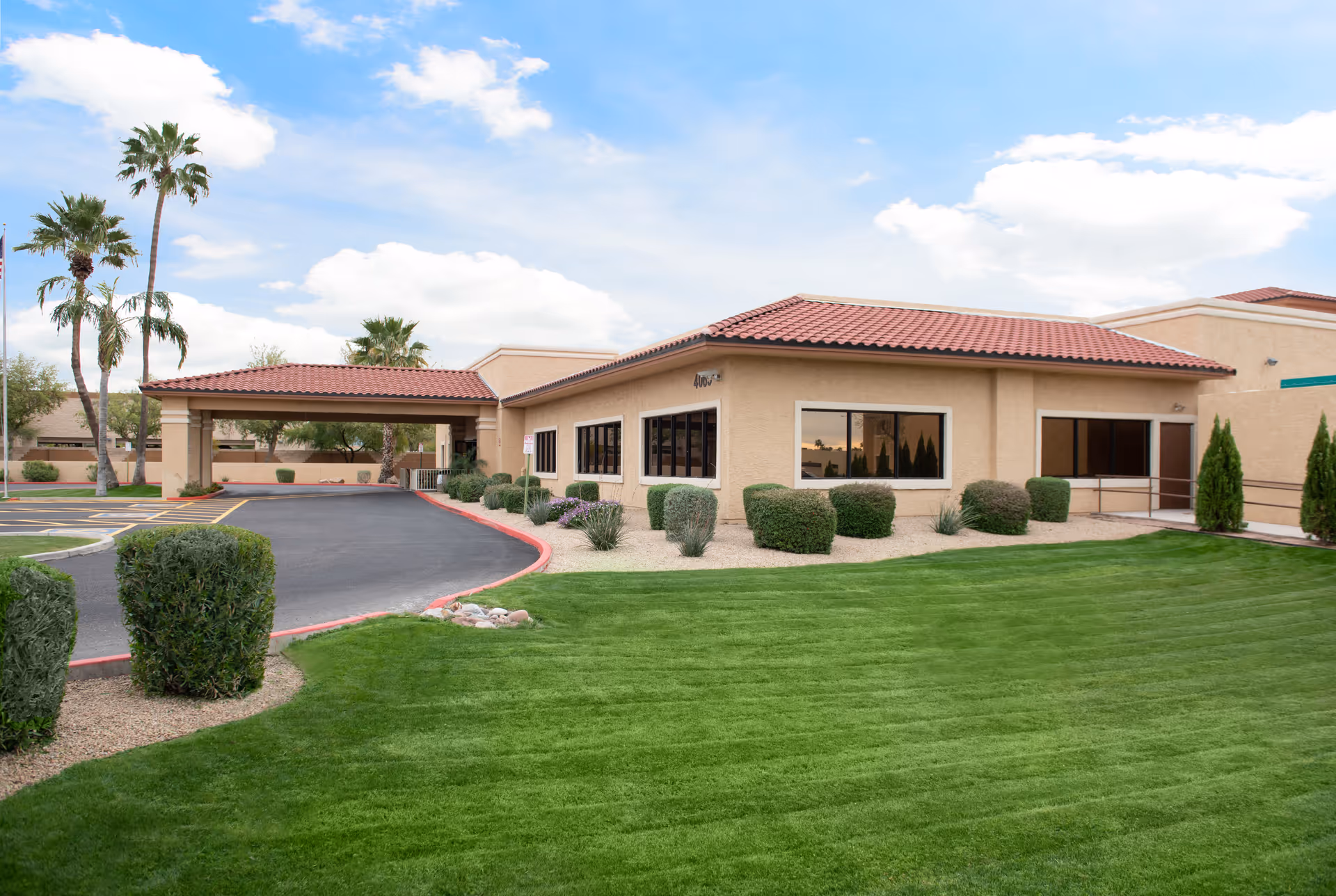 Exterior view of a single-story building with beige stucco walls and a red tile roof, surrounded by neatly trimmed bushes and green grass under a partly cloudy sky. There is a covered driveway entrance and several windows along the building.