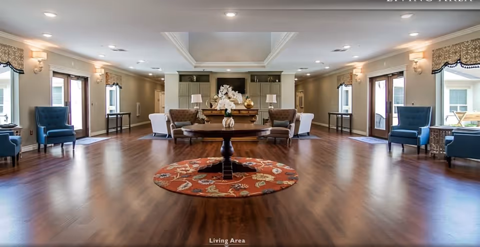 Spacious living area with wooden flooring, a round table with a floral centerpiece on a red patterned rug in the center, surrounded by multiple armchairs in blue and beige colors, wall sconces with lampshades, large windows with valances, and double glass doors leading outside.