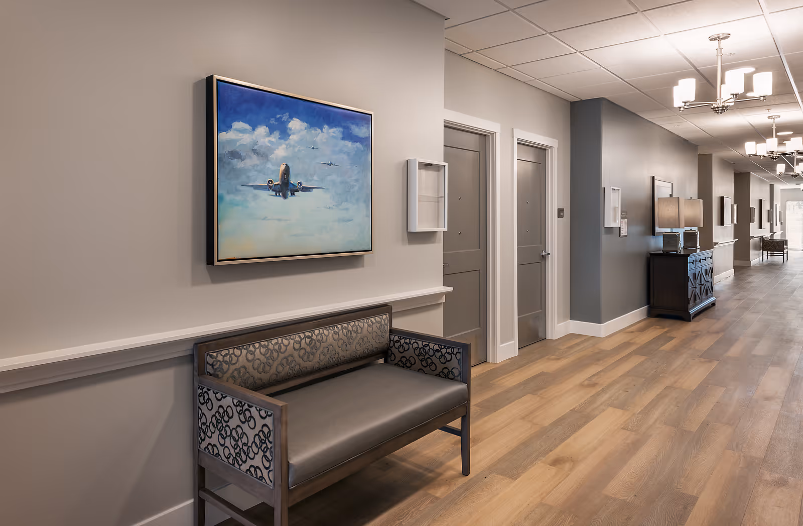 A clean and well-lit hallway in an assisted living facility with light wood flooring, gray walls, and white trim. There is a bench with patterned upholstery along the wall, and a framed painting of an airplane flying in a cloudy sky hangs above it. Several closed doors line the hallway, and a dark wooden cabinet with a lamp is visible further down the corridor. Modern ceiling lights illuminate the space.