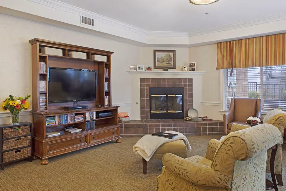 Cozy living room with a wooden entertainment center and TV, a fireplace, armchairs, ottoman and a window with a striped valance.
