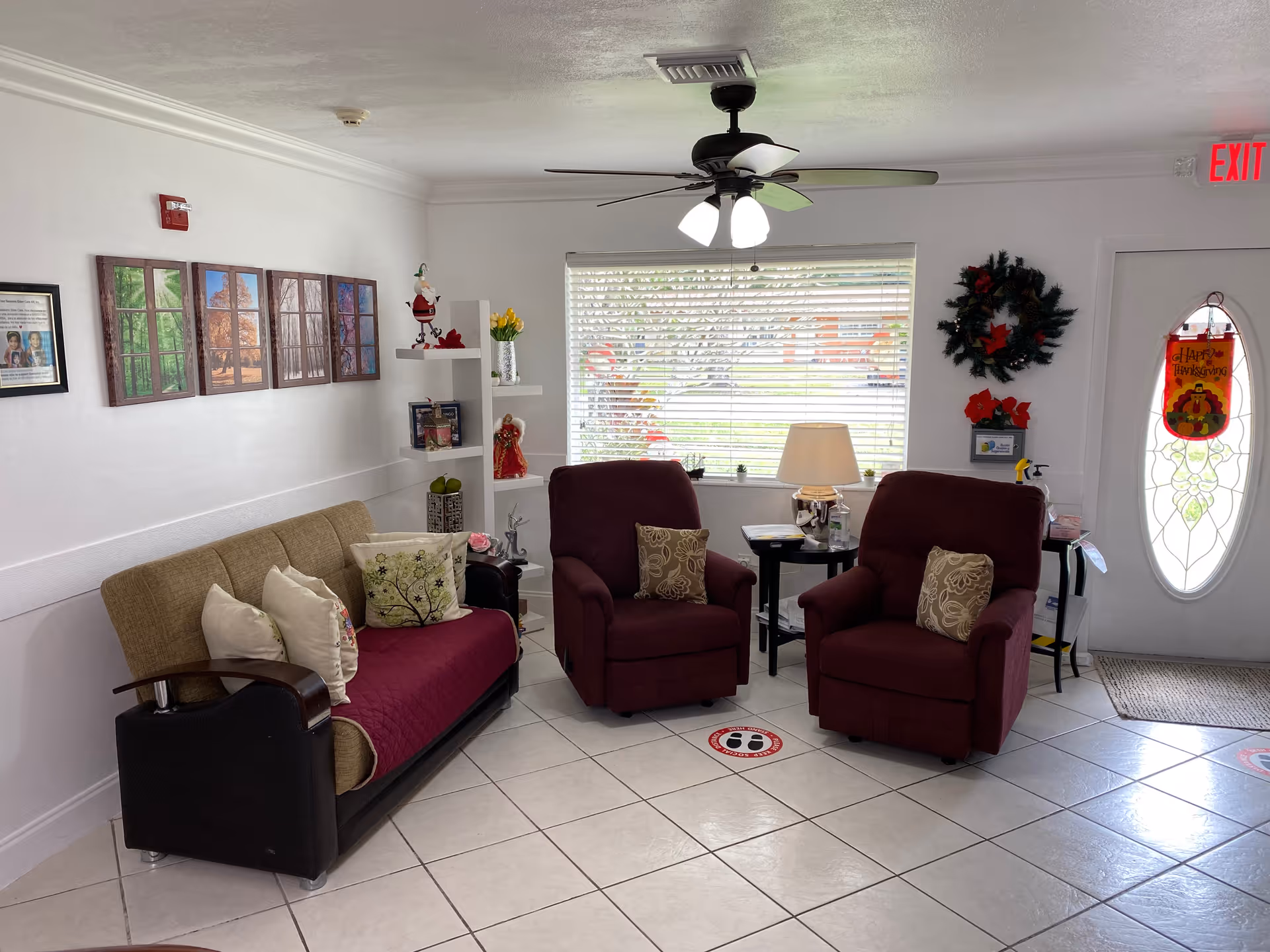 Small communal living room with a sofa, two burgundy recliners, side tables, a ceiling fan, window blinds and holiday decorations.