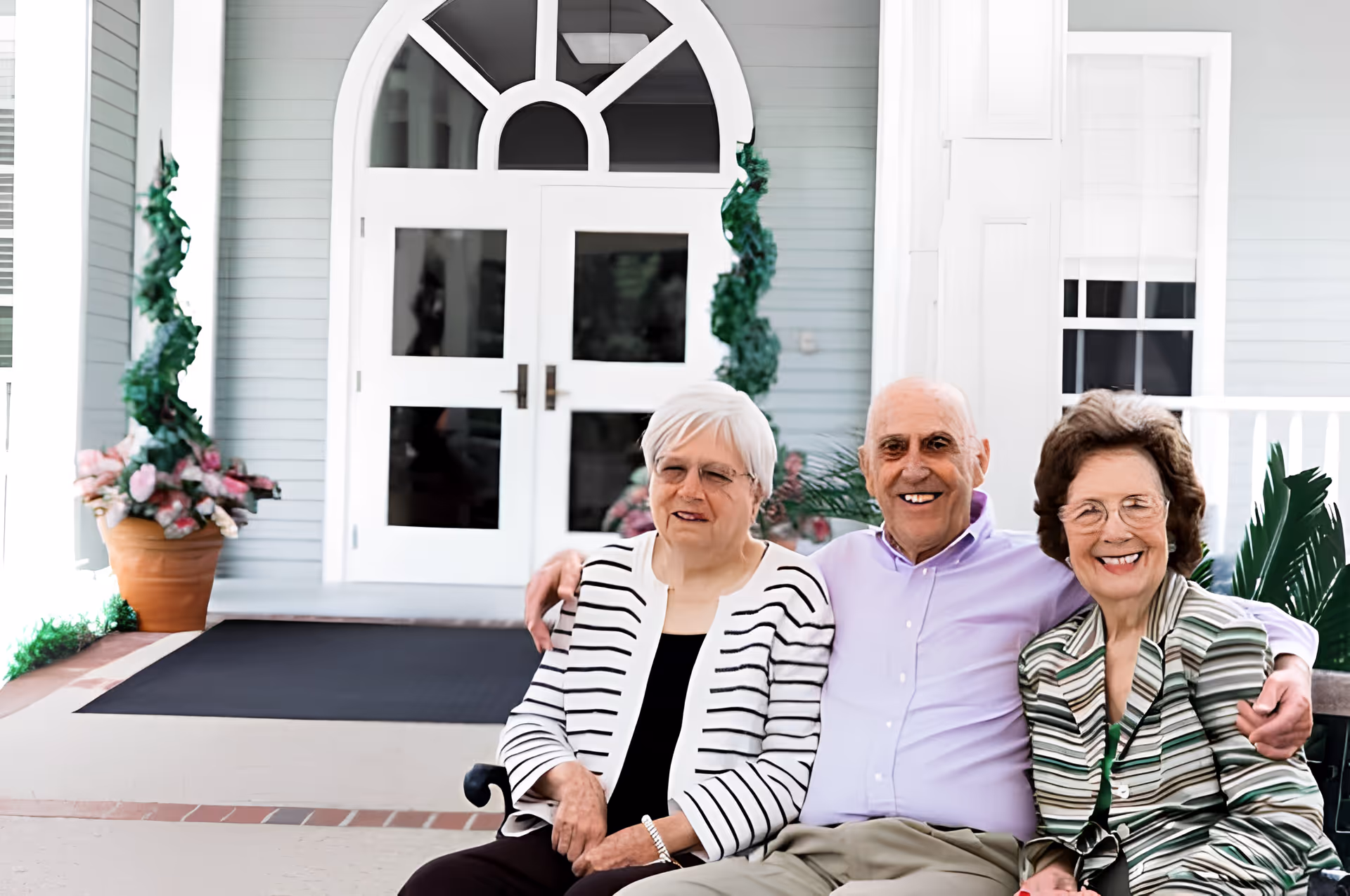 Three elderly individuals sitting closely together on a bench outside a building entrance with white double doors and decorative plants on either side. They are smiling and appear to be enjoying each other's company.
