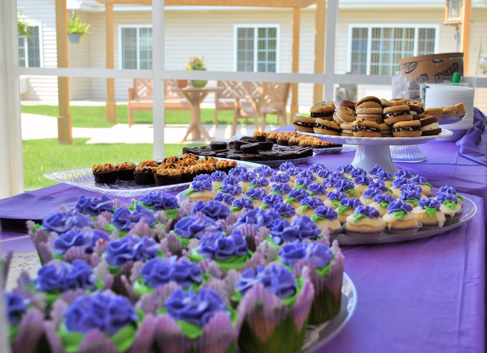 A table covered with a purple tablecloth displaying an assortment of desserts including cupcakes with purple frosting, mini sandwiches, and brownies. The table is set indoors near large windows that show an outdoor patio with chairs and a table.