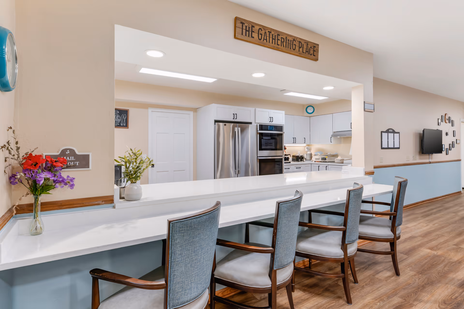 Communal kitchen and serving counter with four chairs, stainless steel appliances, and a sign reading 'The Gathering Place'.