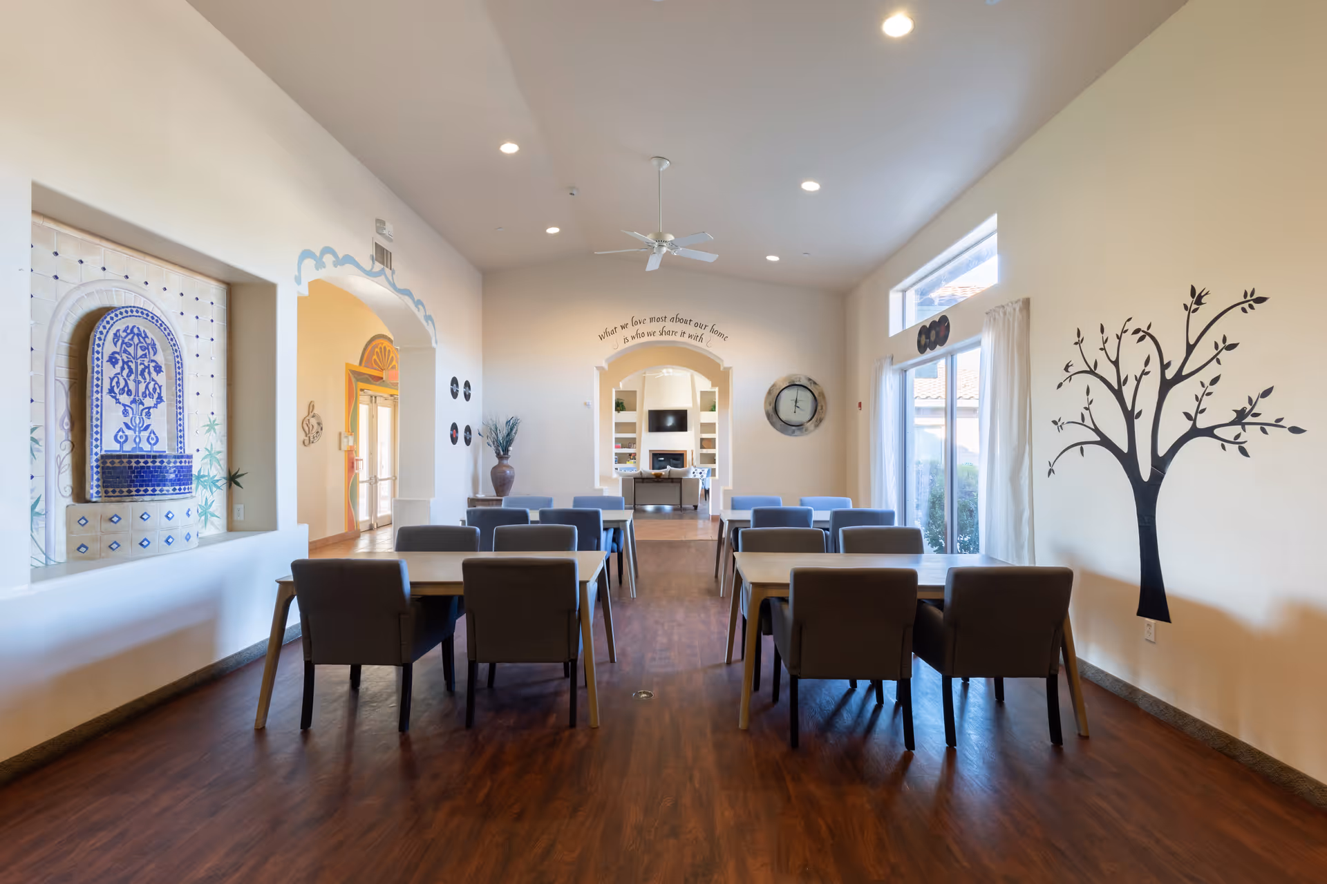 A bright and spacious dining room with several tables and chairs arranged neatly on a wooden floor. The room features a decorative blue and white tiled wall fountain on the left, a large wall clock, and a black tree decal on the right wall. Large windows with curtains allow natural light to fill the space, and an archway leads to another room with shelves and a television. The ceiling has recessed lighting and a ceiling fan.