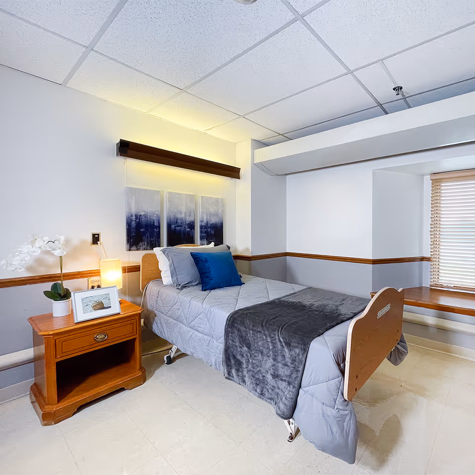 A tidy single-bed patient room with a nightstand, lamp, wall art, and window bench in a senior care facility.