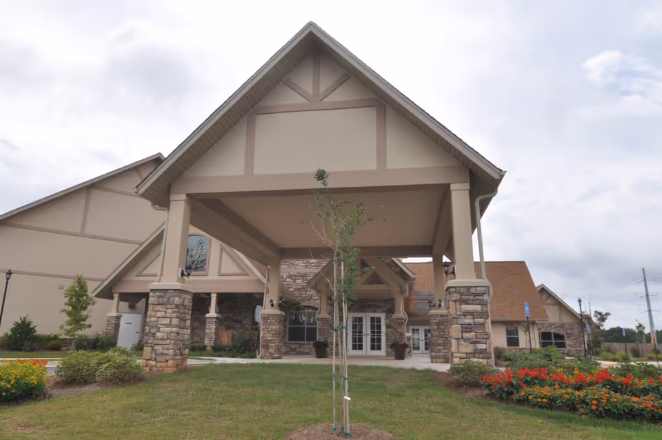 Front entrance of a senior living facility featuring a covered porte-cochere with stone pillars and landscaped grounds.