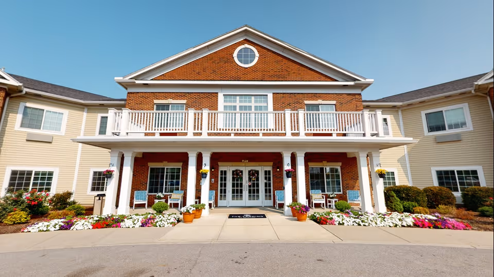 Front entrance of a two-story senior living building with a covered porch, white columns, balcony, seating and colorful flowerbeds.
