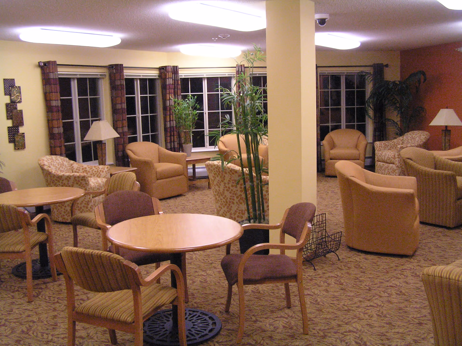 A cozy common area with multiple armchairs and small round tables surrounded by chairs. The room has large windows with patterned curtains, several potted plants, and warm lighting from ceiling fixtures and table lamps. The carpet has a leaf pattern, and the walls are painted in soft yellow and orange tones.
