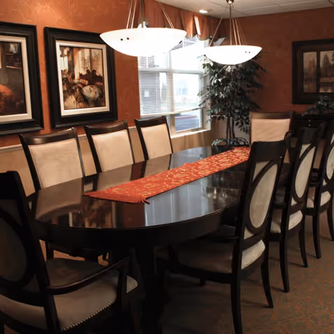 A formal dining room with a long dark wooden table surrounded by cushioned chairs. The table has a red decorative runner. Two pendant lights hang above the table. The walls are decorated with framed artwork and a window with blinds and a valance lets in natural light. A potted plant is placed near the window.
