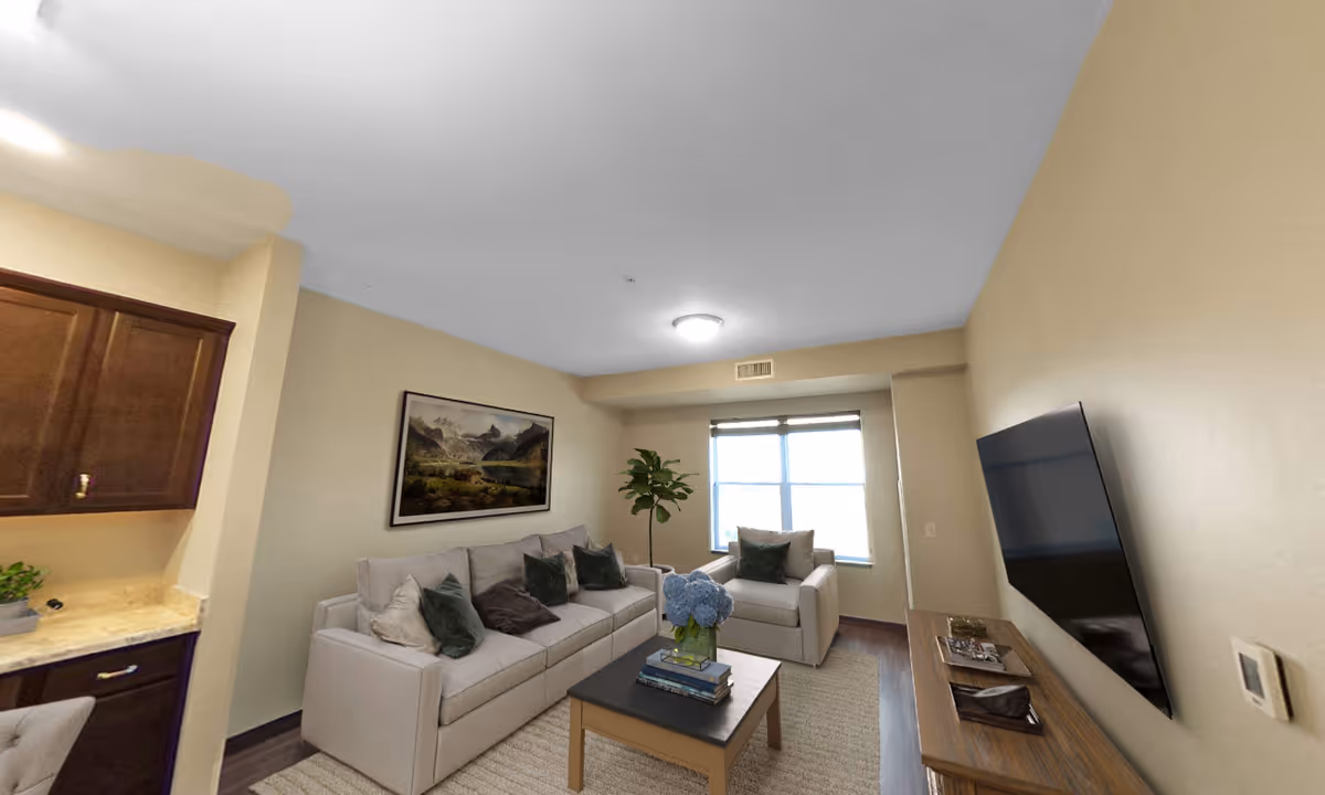 A cozy living room with a beige sofa and armchair adorned with green and brown cushions. A wooden coffee table with a vase of blue flowers and books sits on a light-colored rug. A large flat-screen TV is mounted on the wall opposite the sofa. There is a window with blinds letting in natural light, a potted plant in the corner, and a landscape painting on the wall above the sofa. Part of a kitchen counter with dark wooden cabinets is visible on the left side.