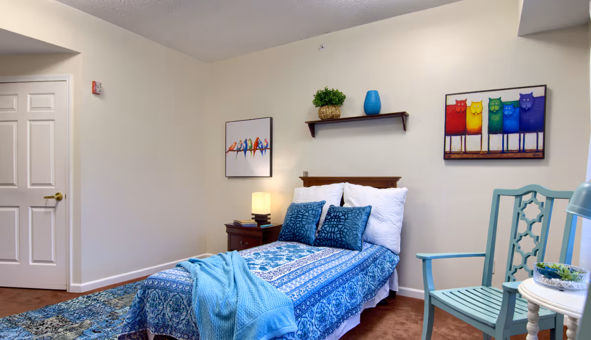 A cozy bedroom with a single bed covered in blue patterned bedding and two blue decorative pillows. There is a wooden nightstand with a lamp and books next to the bed. On the wall above the bed, there is a small shelf with a green plant and a blue vase. Two colorful paintings featuring birds and cats hang on the walls. A light blue wooden chair and a small white table with a plant are positioned near the bed. The room has beige walls and a white door with a gold handle.