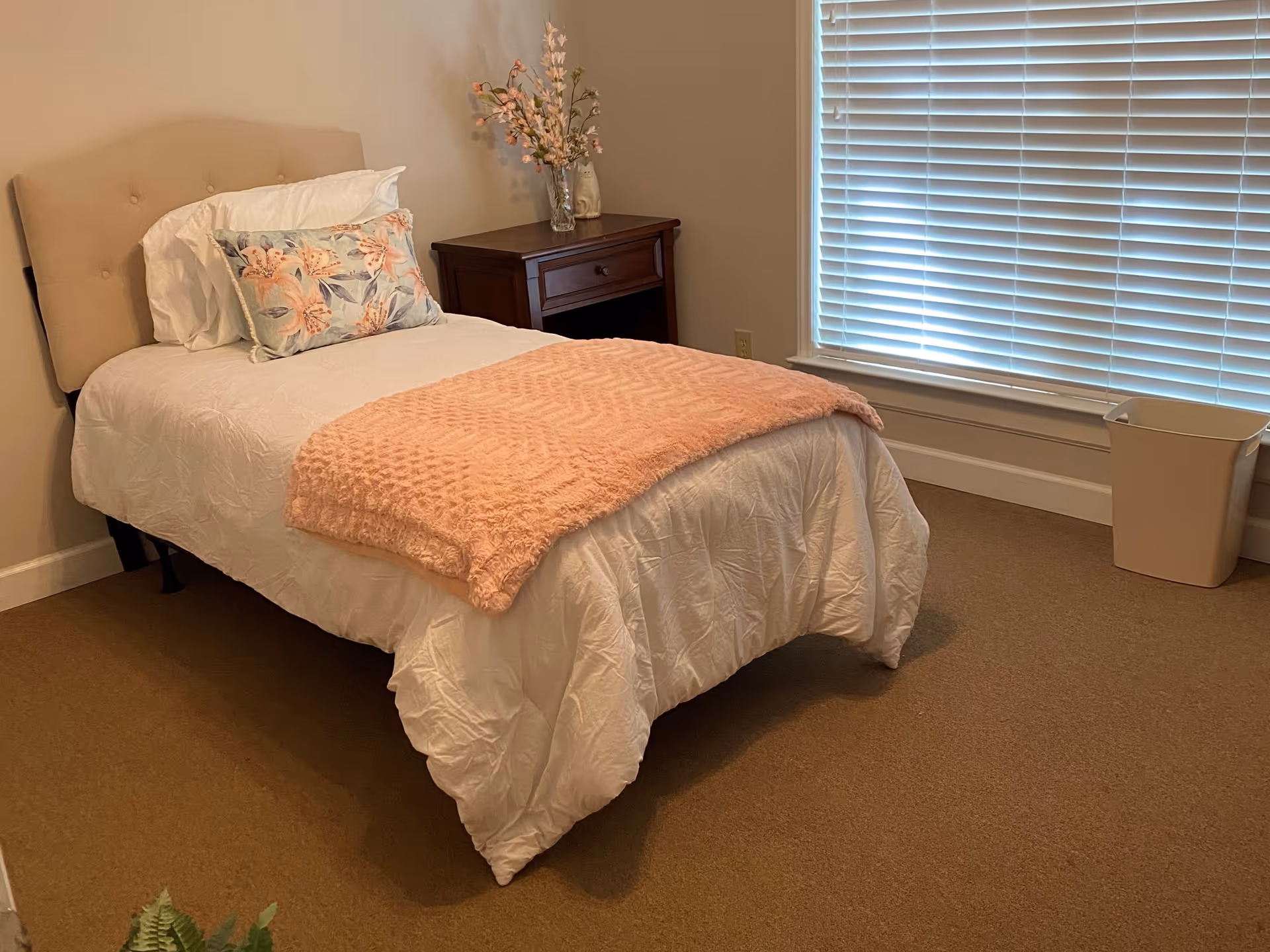 Single bedroom with a neatly made twin bed dressed in white linens, a pink throw and floral pillow, a wooden nightstand with flowers, and a window with closed blinds.