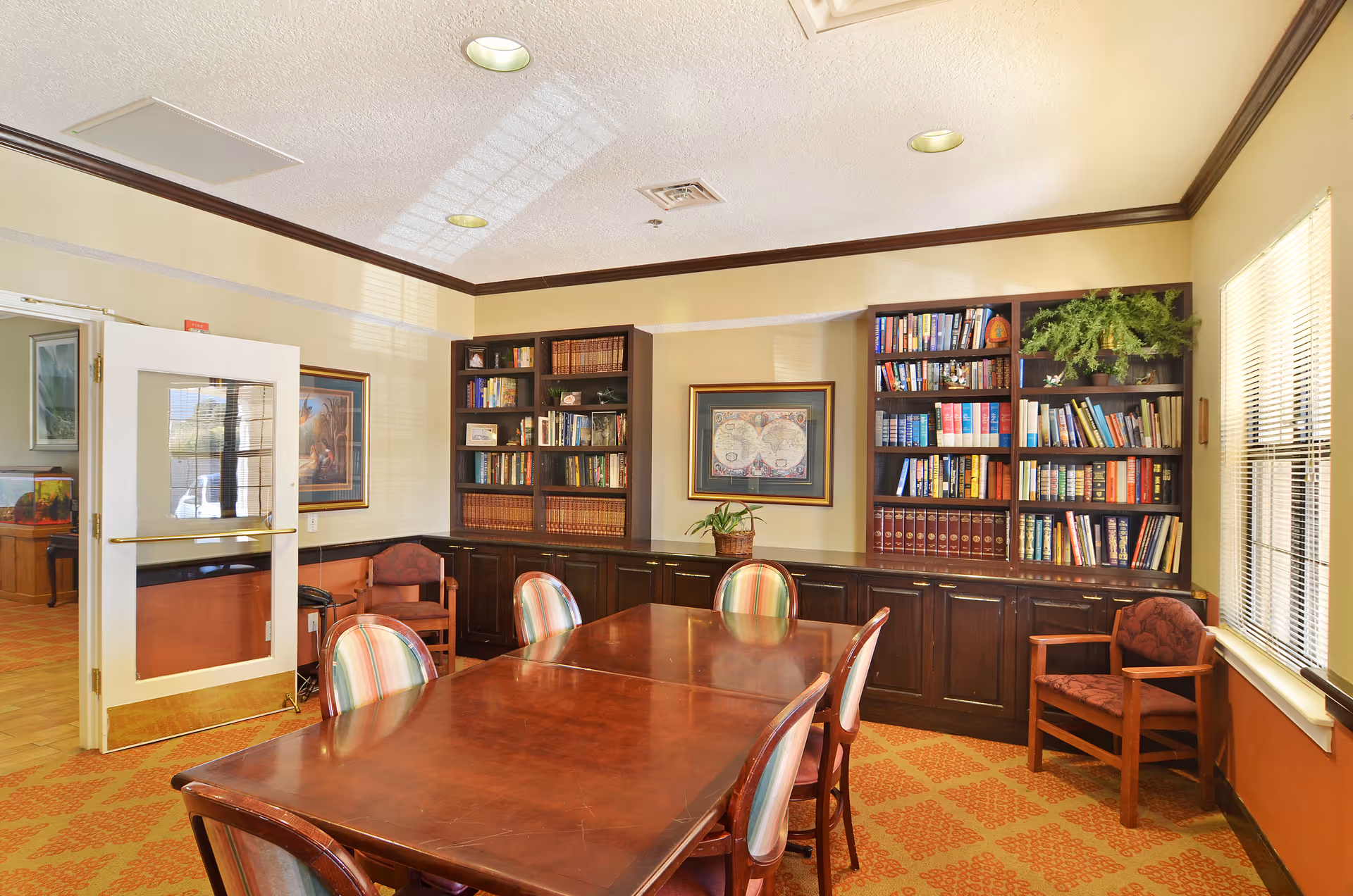 A well-lit room with a wooden table surrounded by six chairs with striped upholstery. The room features built-in dark wood bookshelves filled with books and decorative items, a framed map on the wall, a potted plant, and a window with blinds letting in natural light. The door to the room is open, showing a glimpse of an adjacent area.
