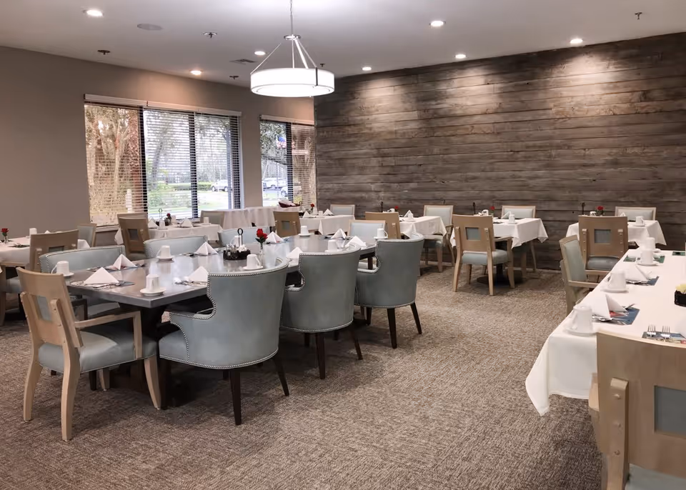 A dining room with multiple tables covered with white tablecloths, set with white napkins, cups, and silverware. The room has a carpeted floor, a wooden accent wall, large windows with blinds, and a modern circular ceiling light fixture.