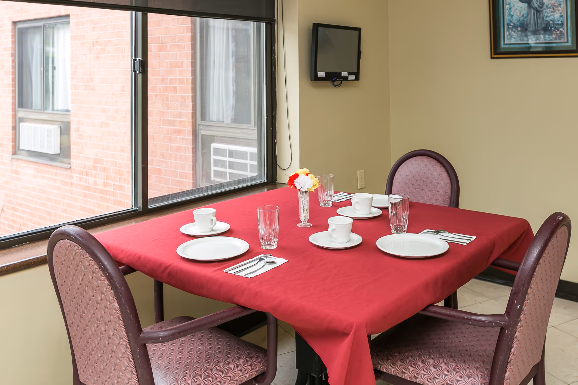 A small dining table set for four with a red tablecloth, plates, cups and a flower centerpiece beside a large window and wall-mounted TV.