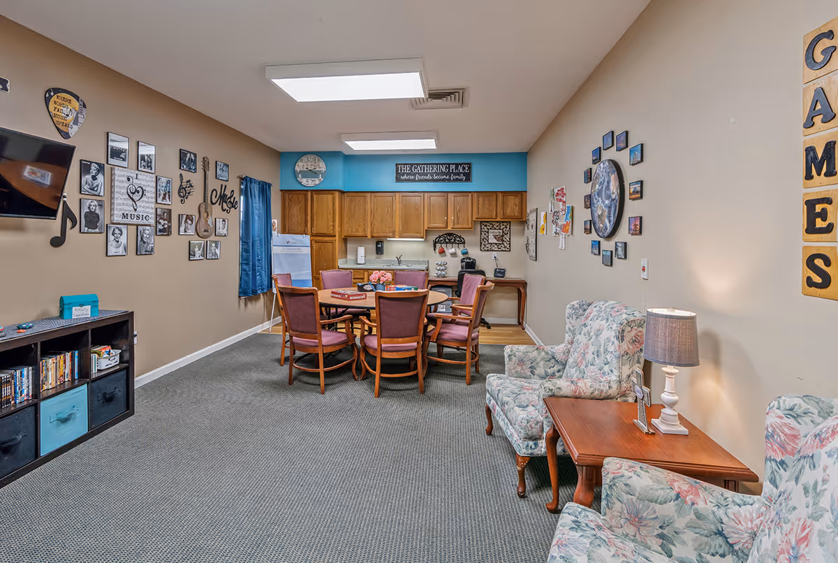 A senior living common room with a round table and chairs, a kitchenette in the back, and floral armchairs beside a side table.