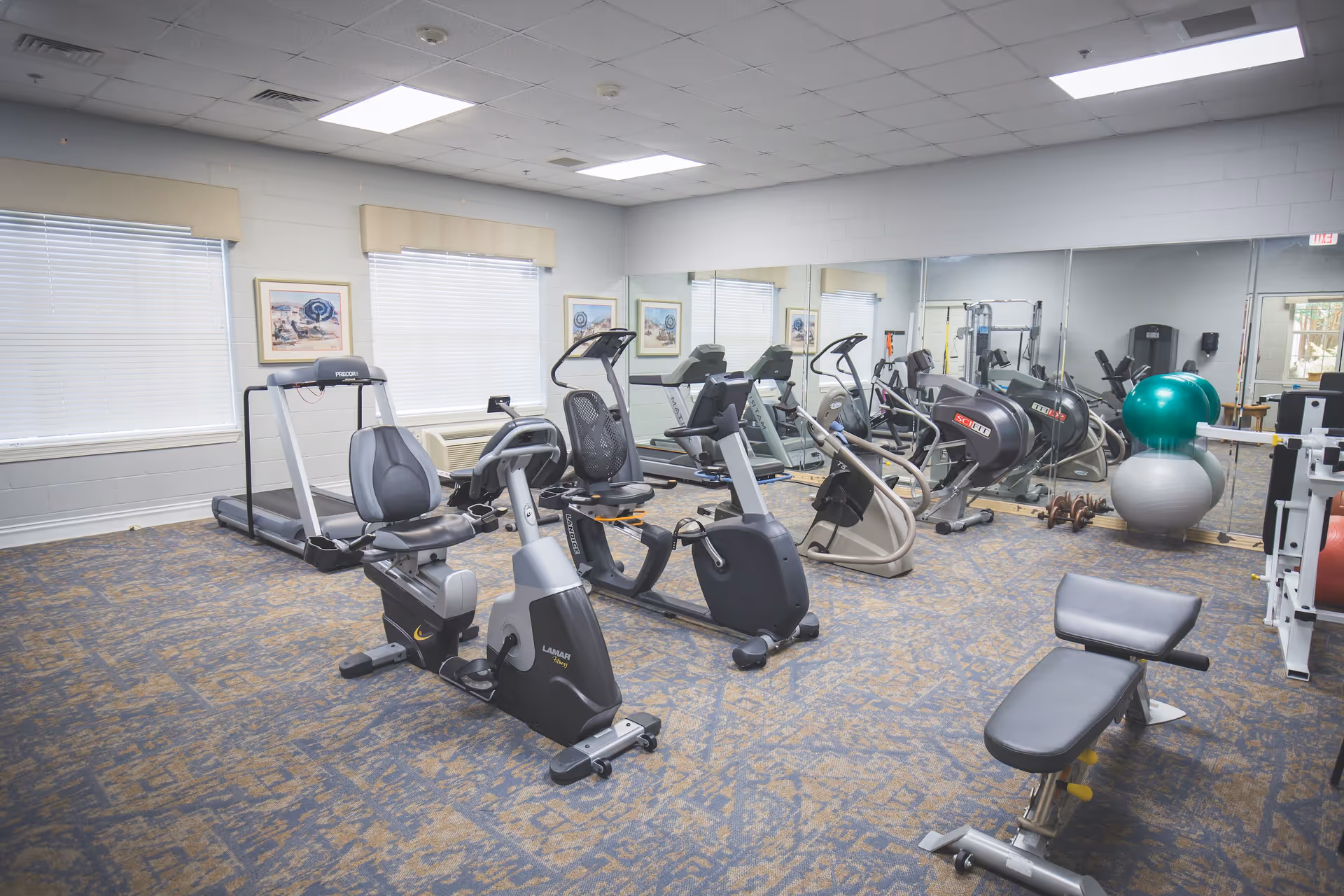 A fitness room in a senior living facility with various exercise equipment including recumbent bikes, treadmills, elliptical machines, a weight bench, and exercise balls. The room has carpeted flooring, large windows with blinds, framed pictures on the walls, and a large mirror covering one wall.
