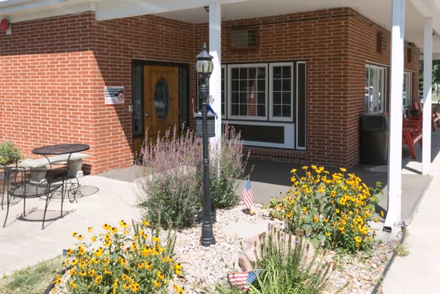 Outdoor view of a brick building entrance with a wooden door, a black lamp post, and flower beds with yellow flowers and small American flags. There is a small round metal table with two chairs on the left side and a covered walkway supported by white pillars.