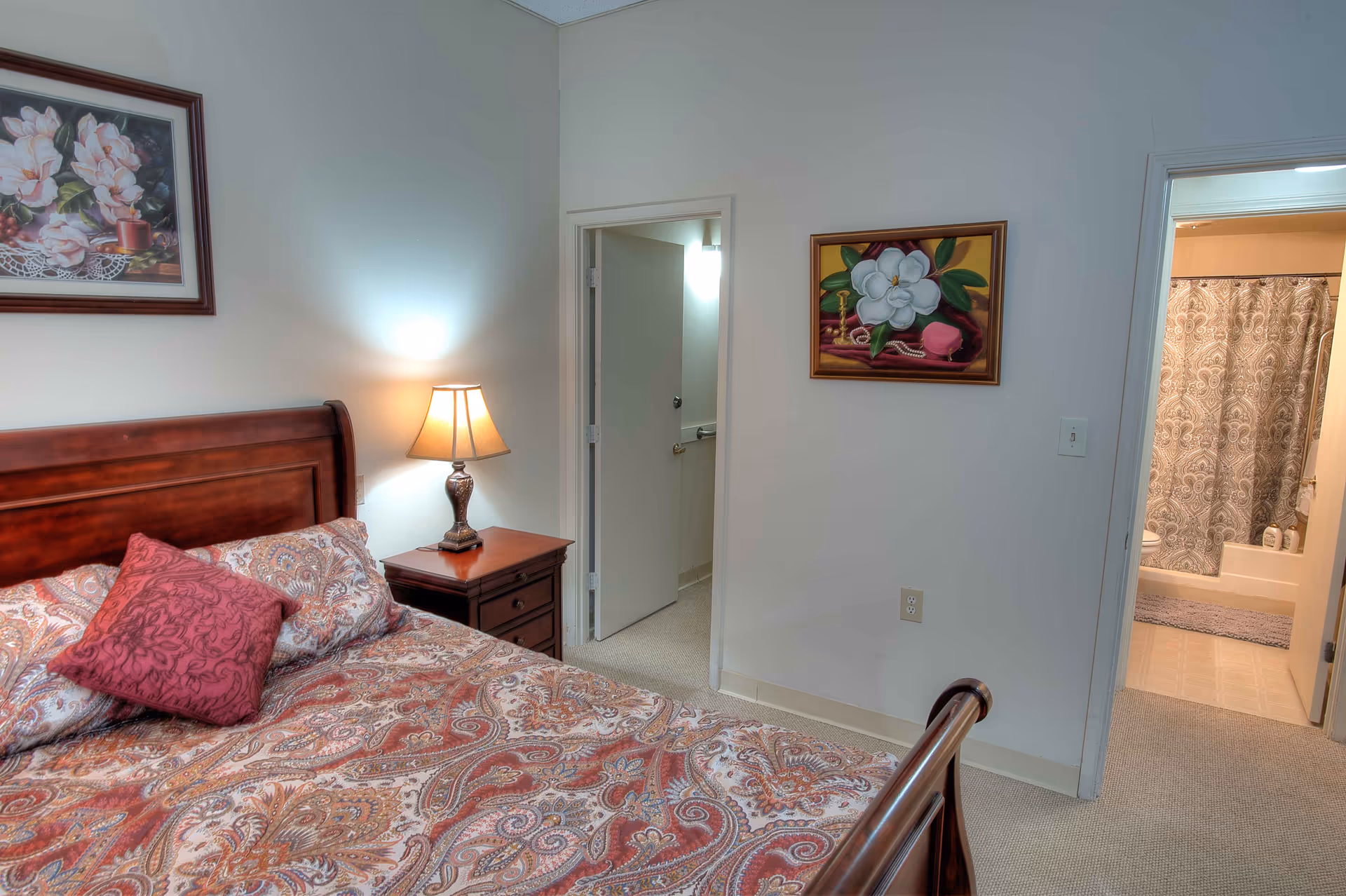 A bedroom with a wooden bed frame and a patterned bedspread in shades of red and beige. There is a nightstand with a lamp next to the bed. Two paintings with floral themes hang on the walls. Two doorways are visible, one leading to a bathroom with a shower curtain and the other to a hallway.