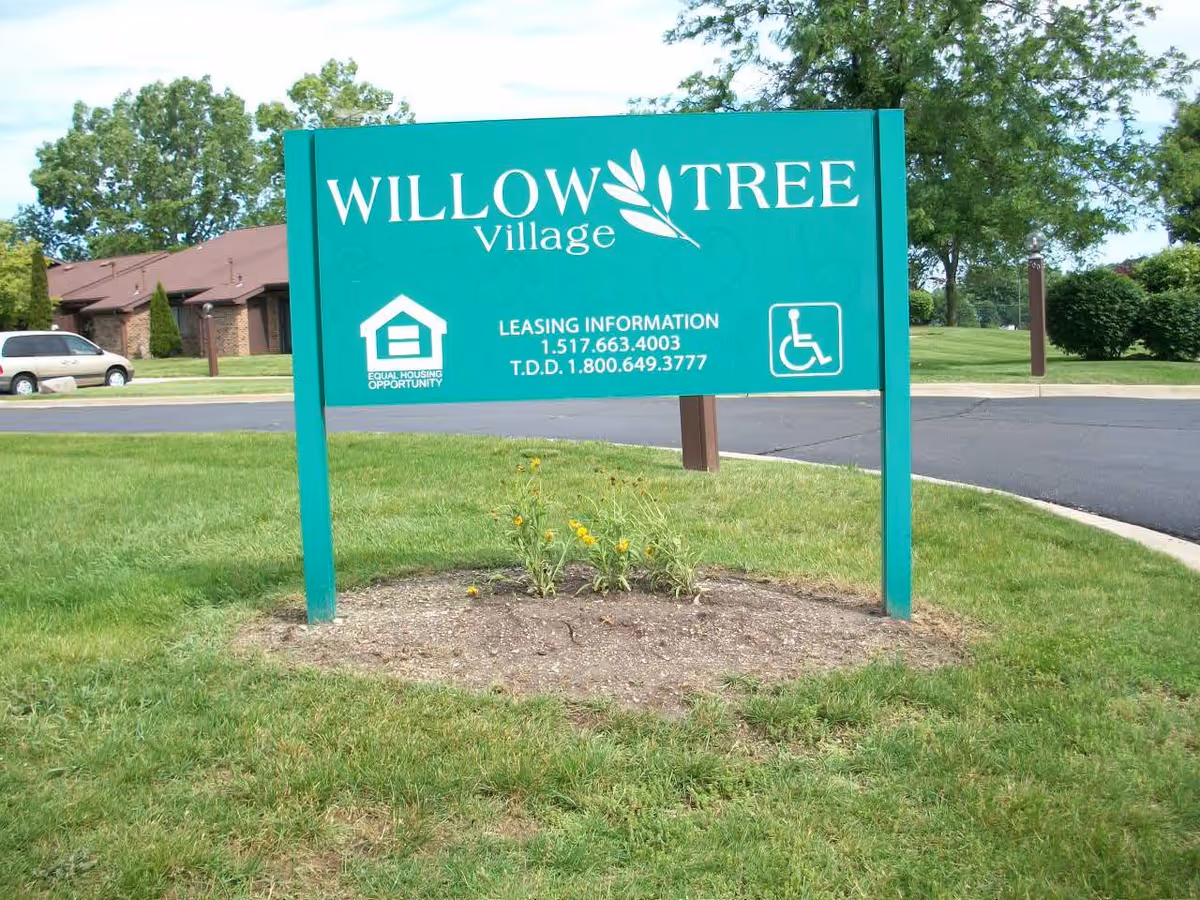 Green sign for Willow Tree Village with leasing information and accessibility symbols, situated on a grassy area near a paved road with trees and buildings in the background.