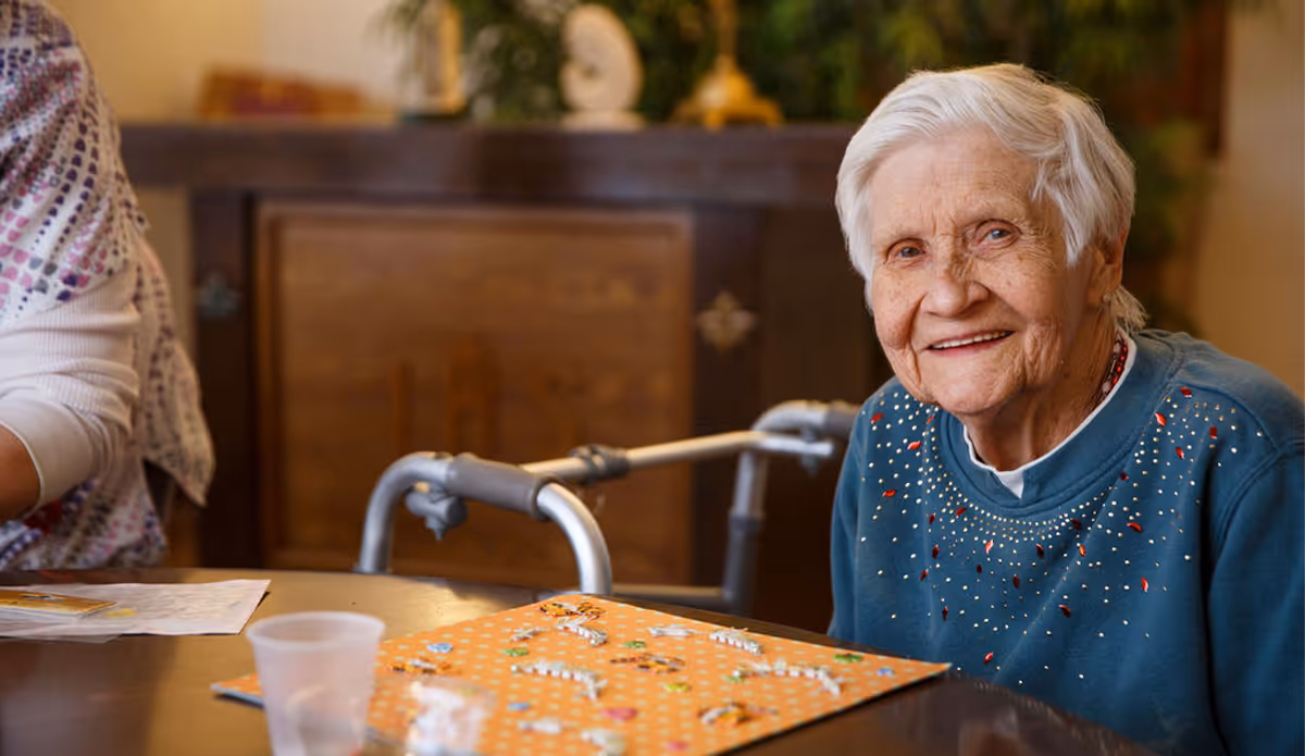 An elderly woman with short white hair wearing a blue sweater with small decorative patterns is sitting at a table, smiling. On the table in front of her is a board game with small pieces. A walker is visible behind her, and part of another person is seen on the left side of the image. The background includes a wooden cabinet and some plants.