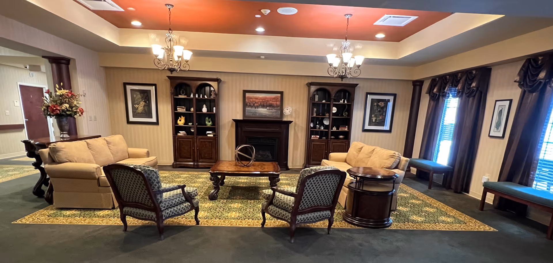A cozy living room area in a senior care facility featuring two beige sofas, two patterned armchairs, a wooden coffee table, and two wooden display cabinets flanking a fireplace. The room has a green and yellow patterned rug, framed artwork on the walls, chandeliers hanging from the ceiling, and windows with dark curtains letting in natural light.