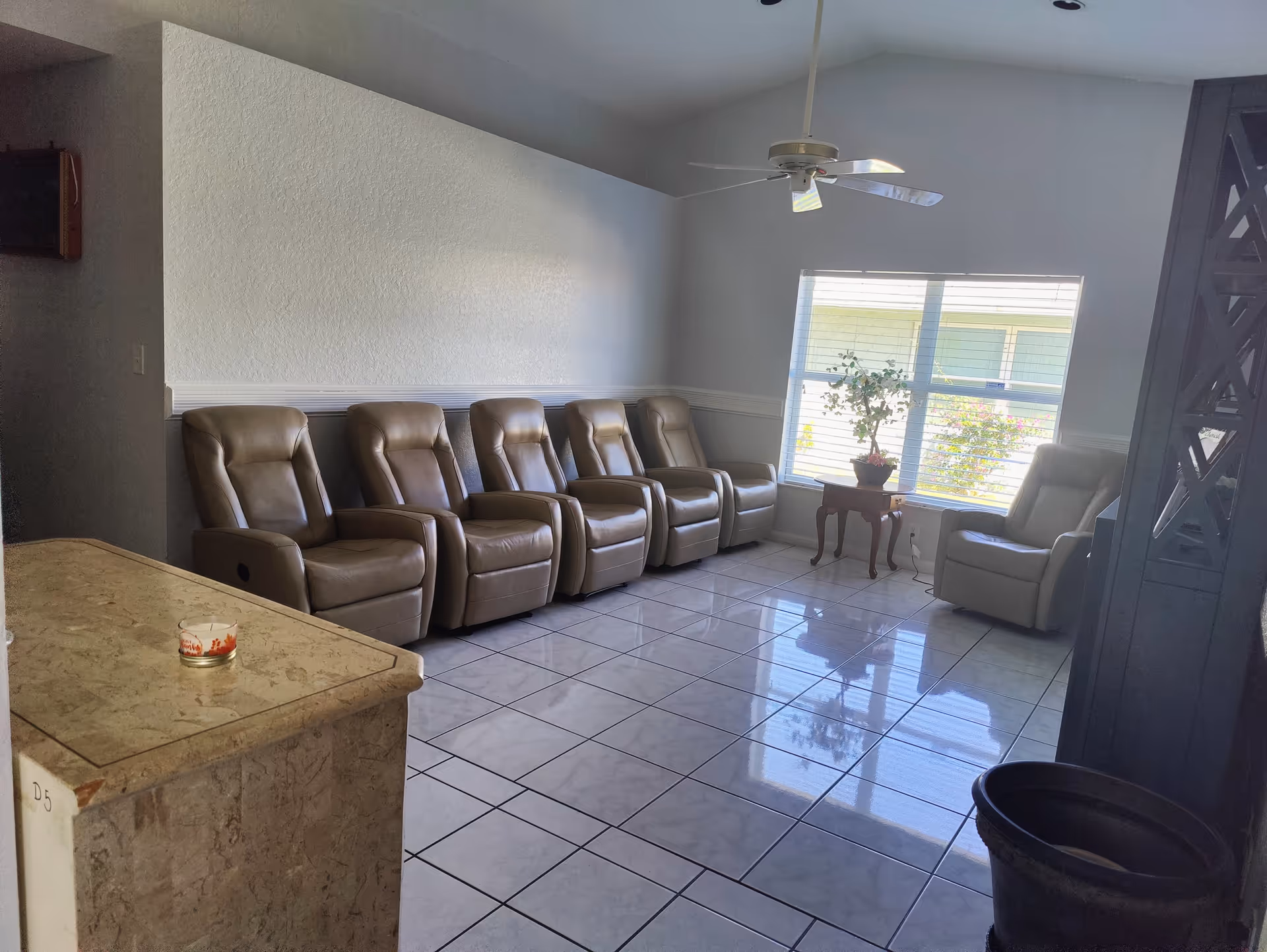 A bright lounge with a row of beige recliners along a wall, a window with a small side table and plant, and tiled floors.