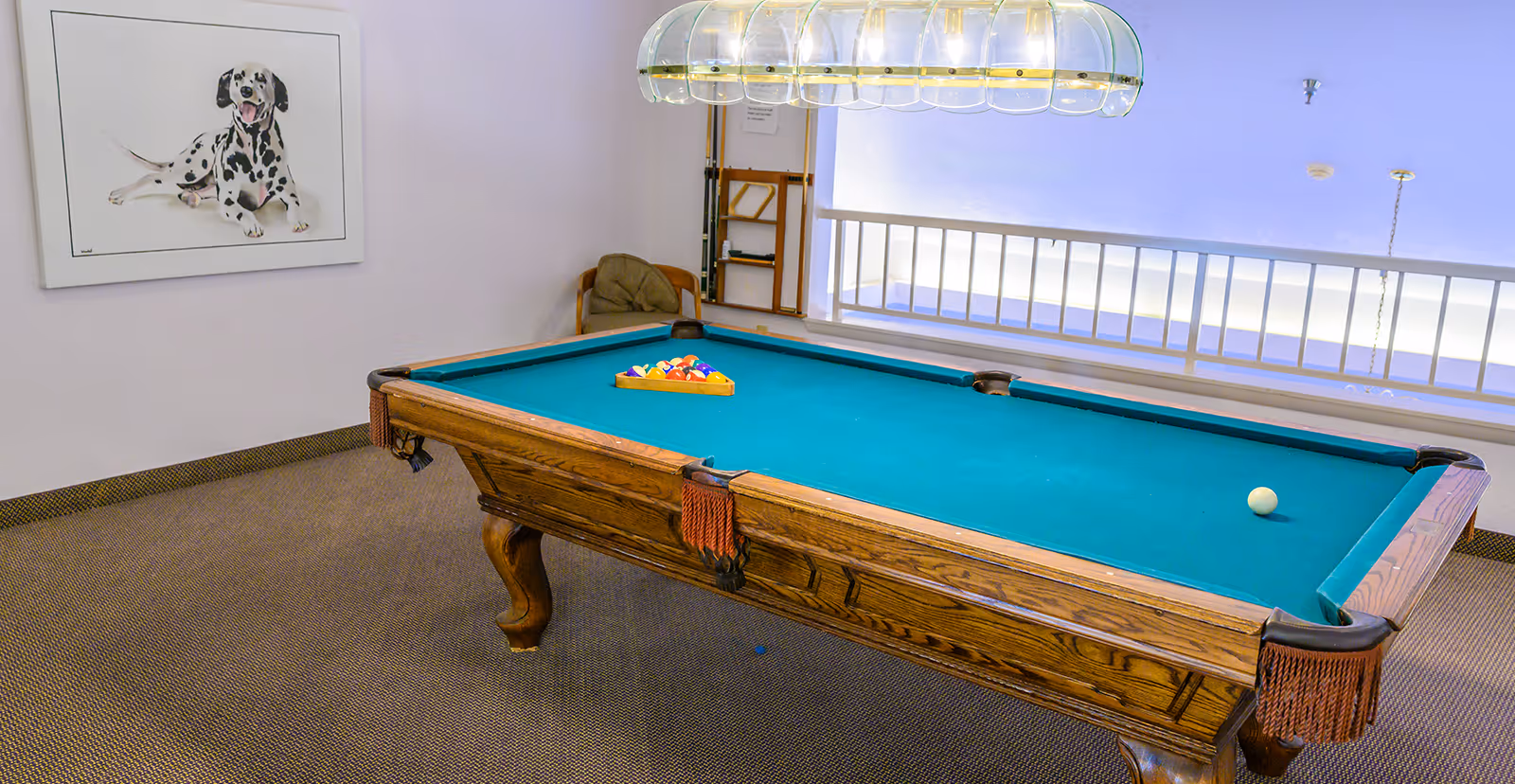 A pool table with a green felt surface and a full rack of billiard balls set up, located in a carpeted room with a framed picture of a Dalmatian dog on the wall and a hanging light fixture above the table.