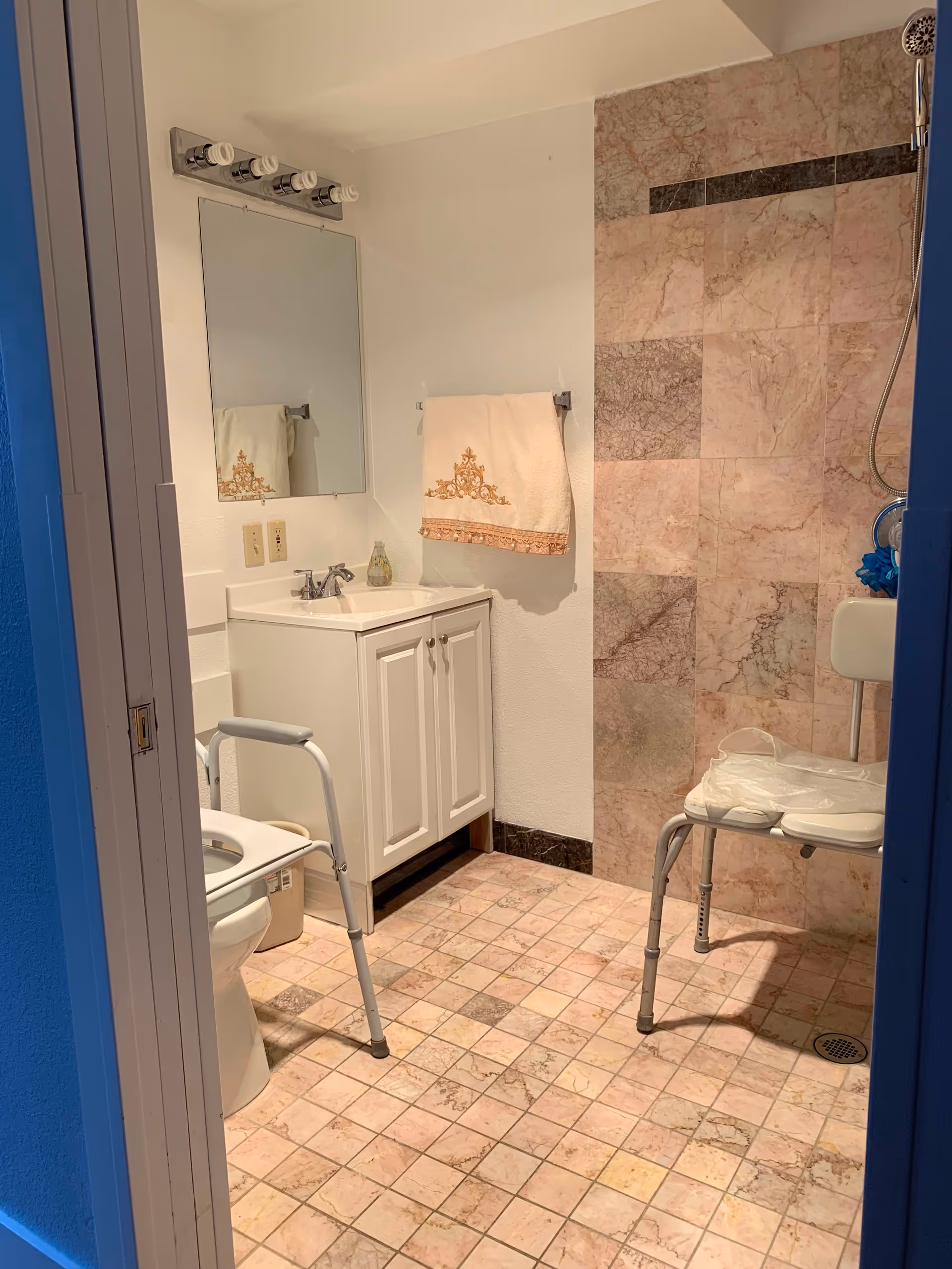 A bathroom with pink and beige tiled floor and wall. There is a white toilet with a safety frame, a white vanity with a sink and a mirror above it, a towel rack with a decorative towel, and a shower area with a white shower chair and a handheld showerhead.