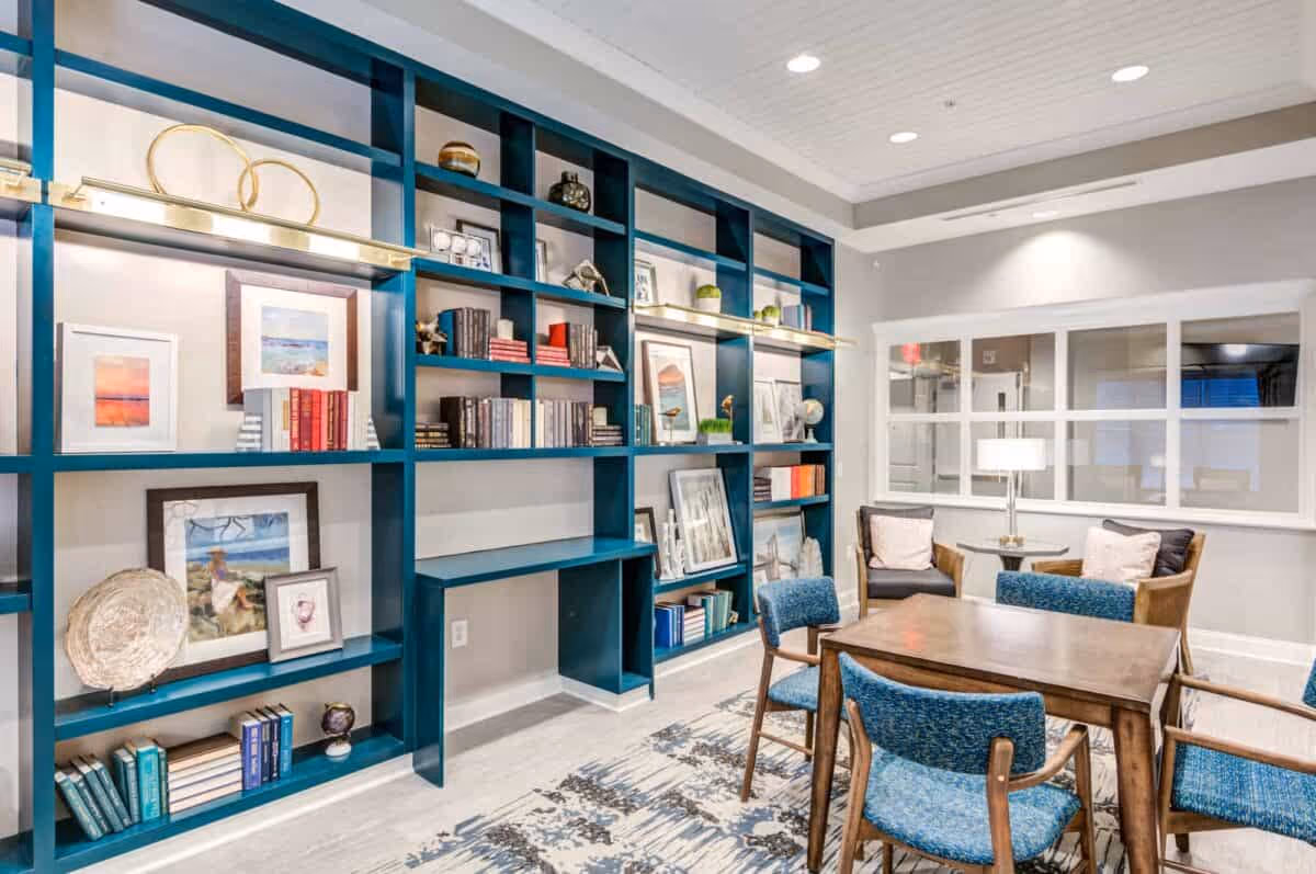 A cozy interior room featuring a large teal bookshelf filled with books, framed pictures, and decorative items. In front of the bookshelf is a wooden table surrounded by four blue upholstered chairs. In the background, there are two armchairs with cushions and a small round table with a lamp. The room has light-colored walls and a patterned rug on the floor.