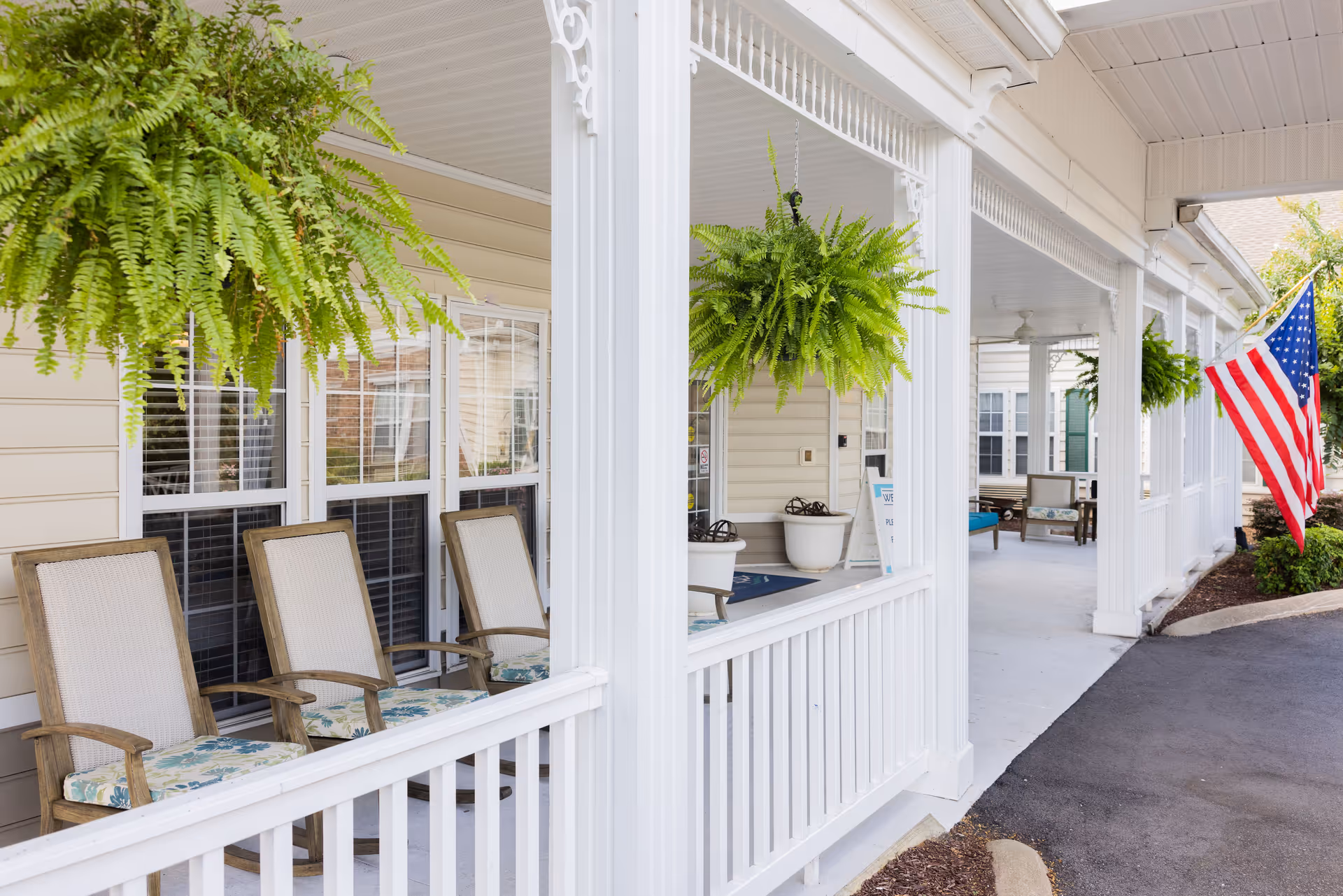 Covered front porch of a senior living facility with hanging ferns, rocking chairs, white columns, and an American flag.