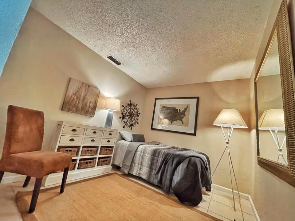 A small, cozy bedroom with a single bed covered in gray bedding. There is a white storage unit with wicker baskets and drawers against one wall, topped with a lamp and decorative items. A brown upholstered chair is positioned near the storage unit. The walls are decorated with framed artwork and a decorative metal piece. A tall floor lamp stands next to a large mirror on the opposite wall. The room has a warm, inviting atmosphere with beige walls and a light-colored floor.