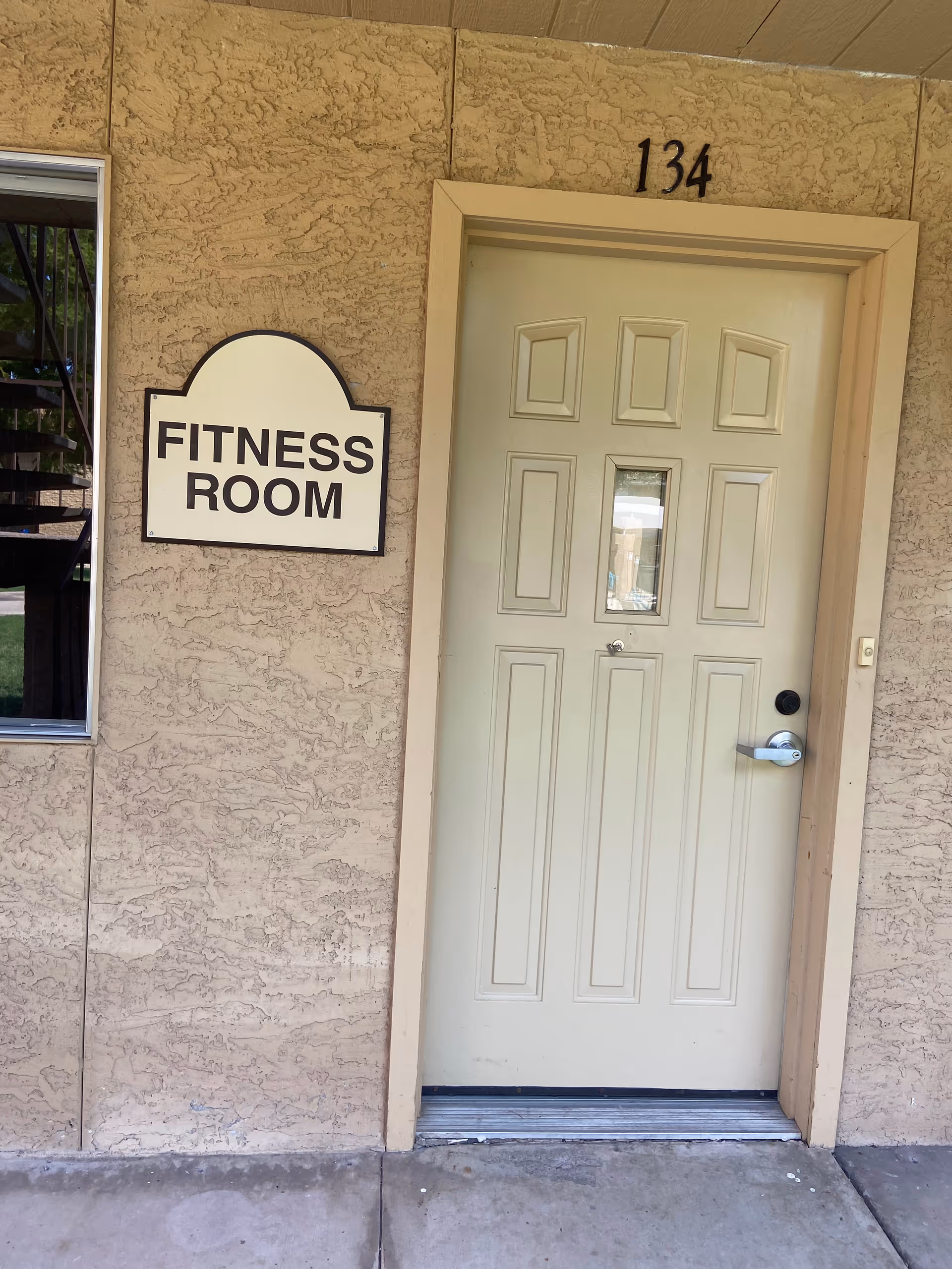 Exterior view of a beige door labeled with the number 134 and a sign next to it that reads 'Fitness Room' on a textured wall.
