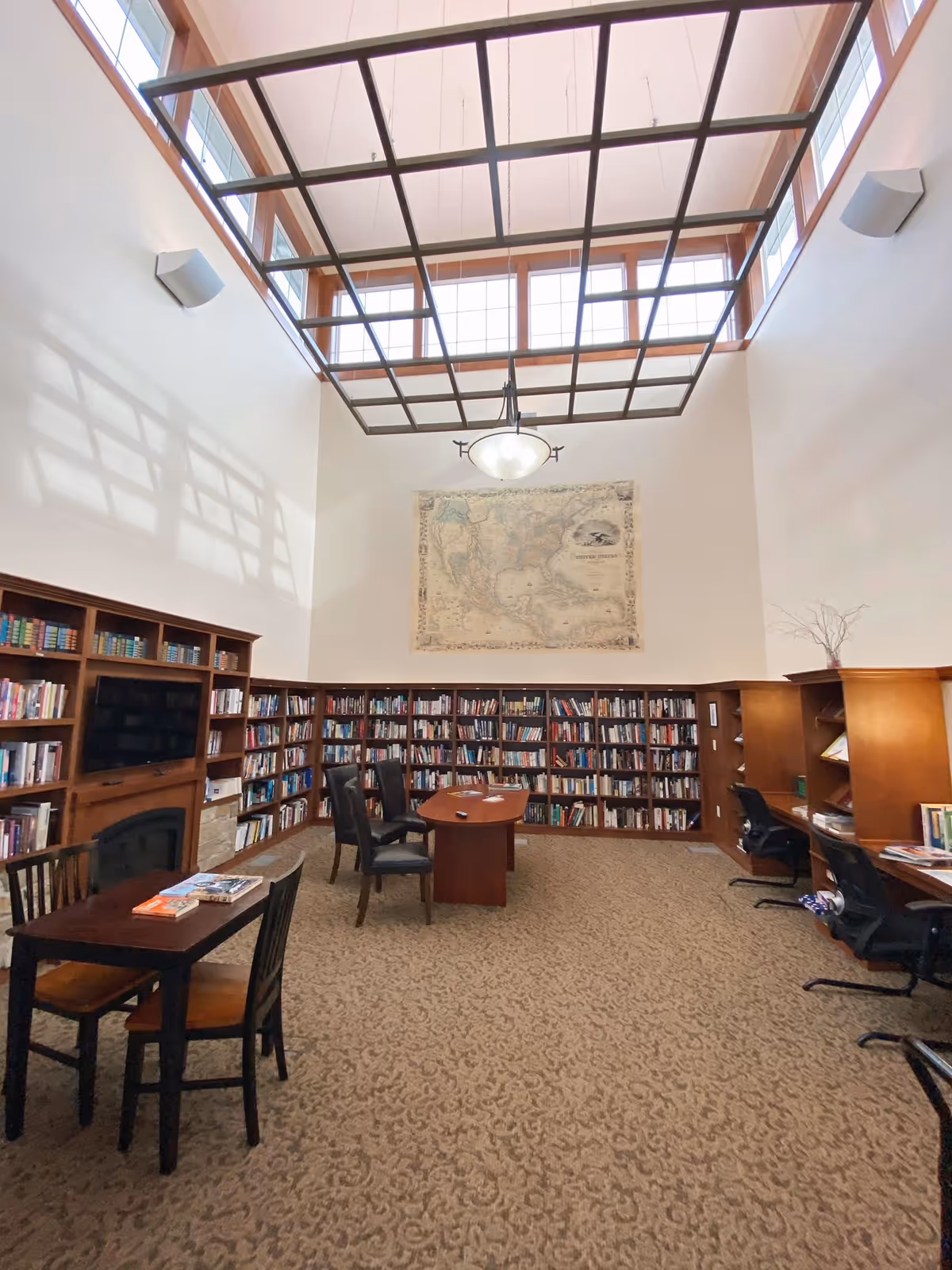 A spacious senior living facility library with high ceilings and large windows near the ceiling. The room features wooden bookshelves filled with books lining the walls, a large map hanging on the far wall, a wooden table with chairs in the center, and a smaller table with chairs in the foreground. There are also desks with office chairs along the right wall. The carpet has a patterned design, and a modern light fixture hangs from the ceiling.