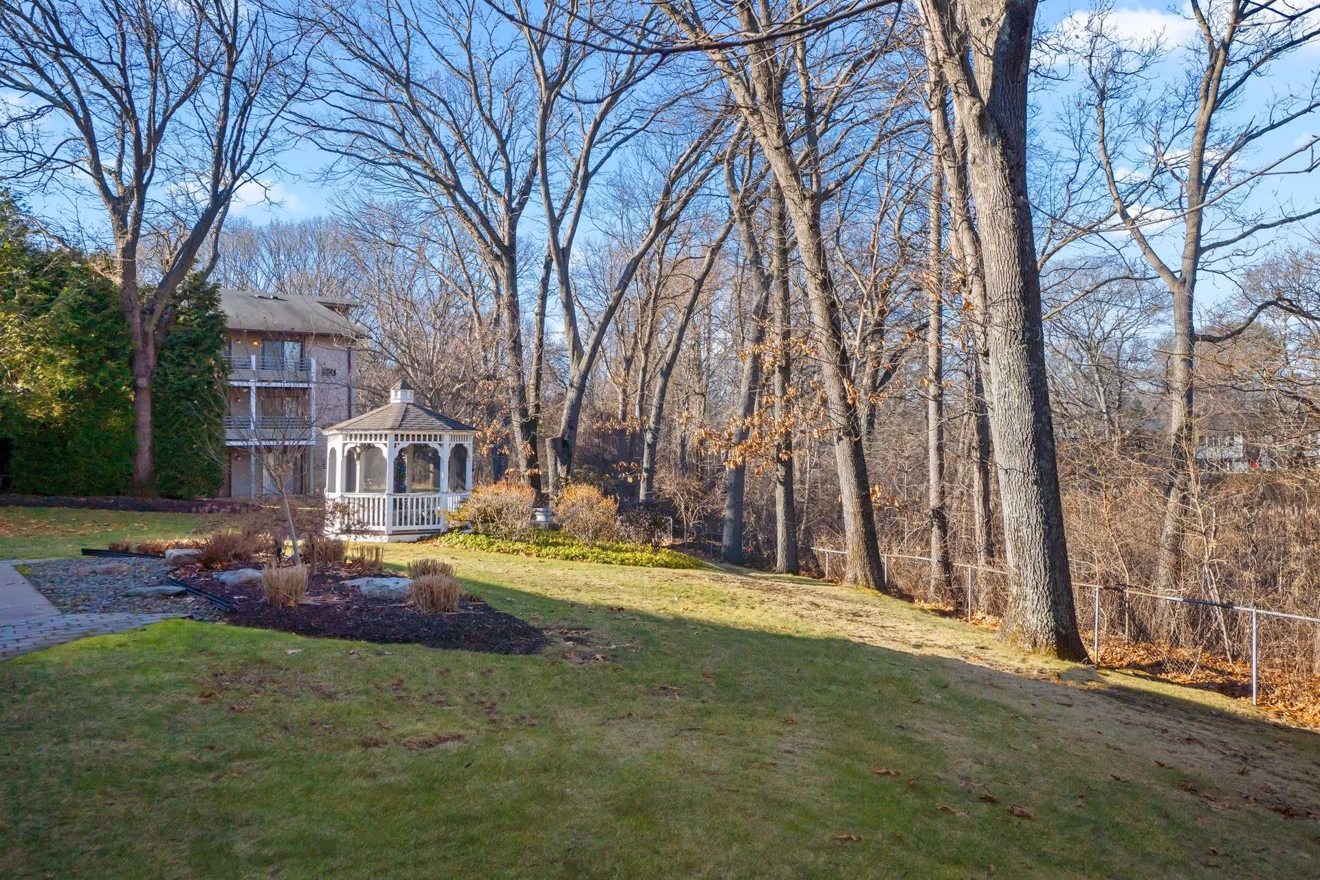 Outdoor garden area with a white gazebo surrounded by trees with bare branches and a grassy lawn. A multi-story building is partially visible on the left side of the image under a clear blue sky.