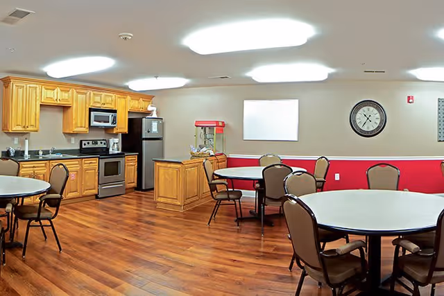A communal kitchen and dining area with wooden cabinets, stainless steel appliances including a microwave, stove, and refrigerator. There are round tables with chairs arranged around the room, a popcorn machine on a small island, a wall clock, and a blank whiteboard on the wall. The floor is wooden and the walls are painted beige with a red lower section.