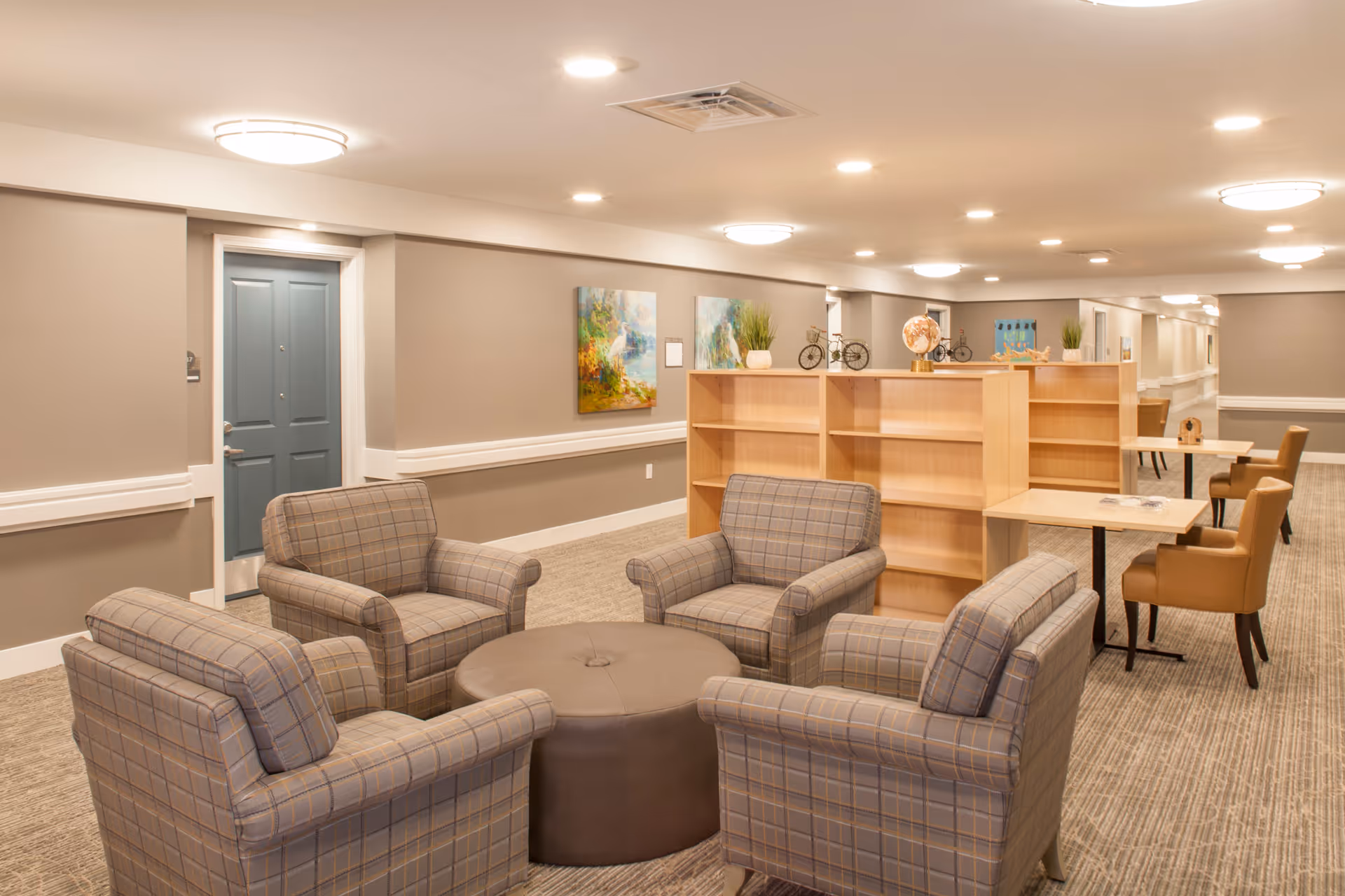 Well-lit common lounge with four upholstered armchairs around a round ottoman, wooden shelving divider and tables with chairs in the background.
