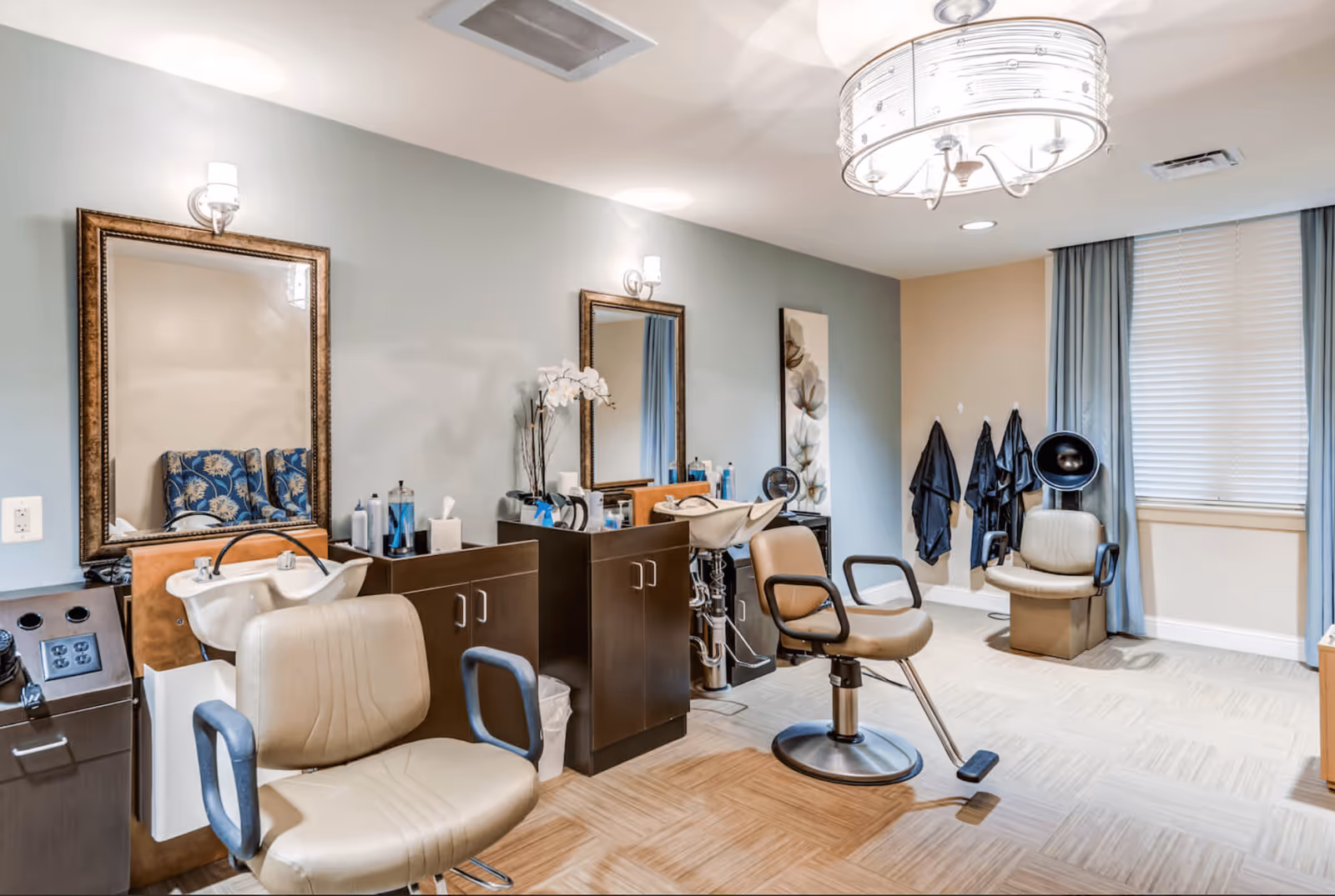 Interior view of a hair salon area in Chantilly Heights Memory Care facility featuring two beige salon chairs with armrests, two wash basins, large framed mirrors on a light blue wall, dark wood cabinets with hair care products, a wall-mounted hair dryer, and a window with blinds and blue curtains.