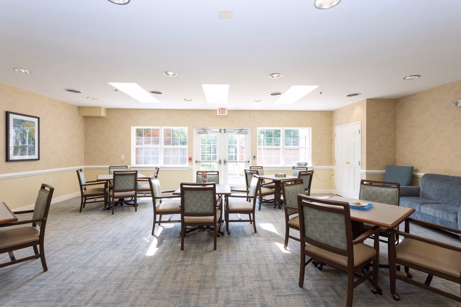 A bright and spacious common area in a senior living facility with multiple tables and chairs arranged for dining or activities. The room has beige walls, large windows letting in natural light, a blue couch, and a framed picture on the wall.