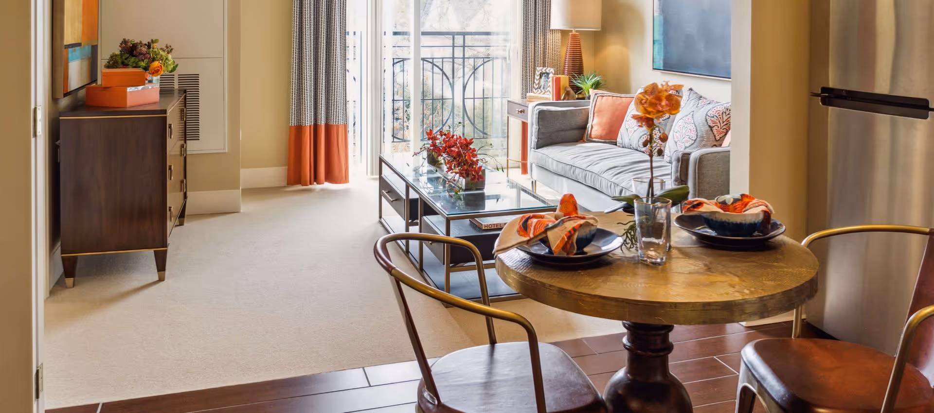 A cozy living room and dining area with a round wooden table set for two, a gray sofa with decorative pillows, a glass coffee table with red flowers, a sideboard with a flower arrangement, and large windows with patterned curtains letting in natural light.