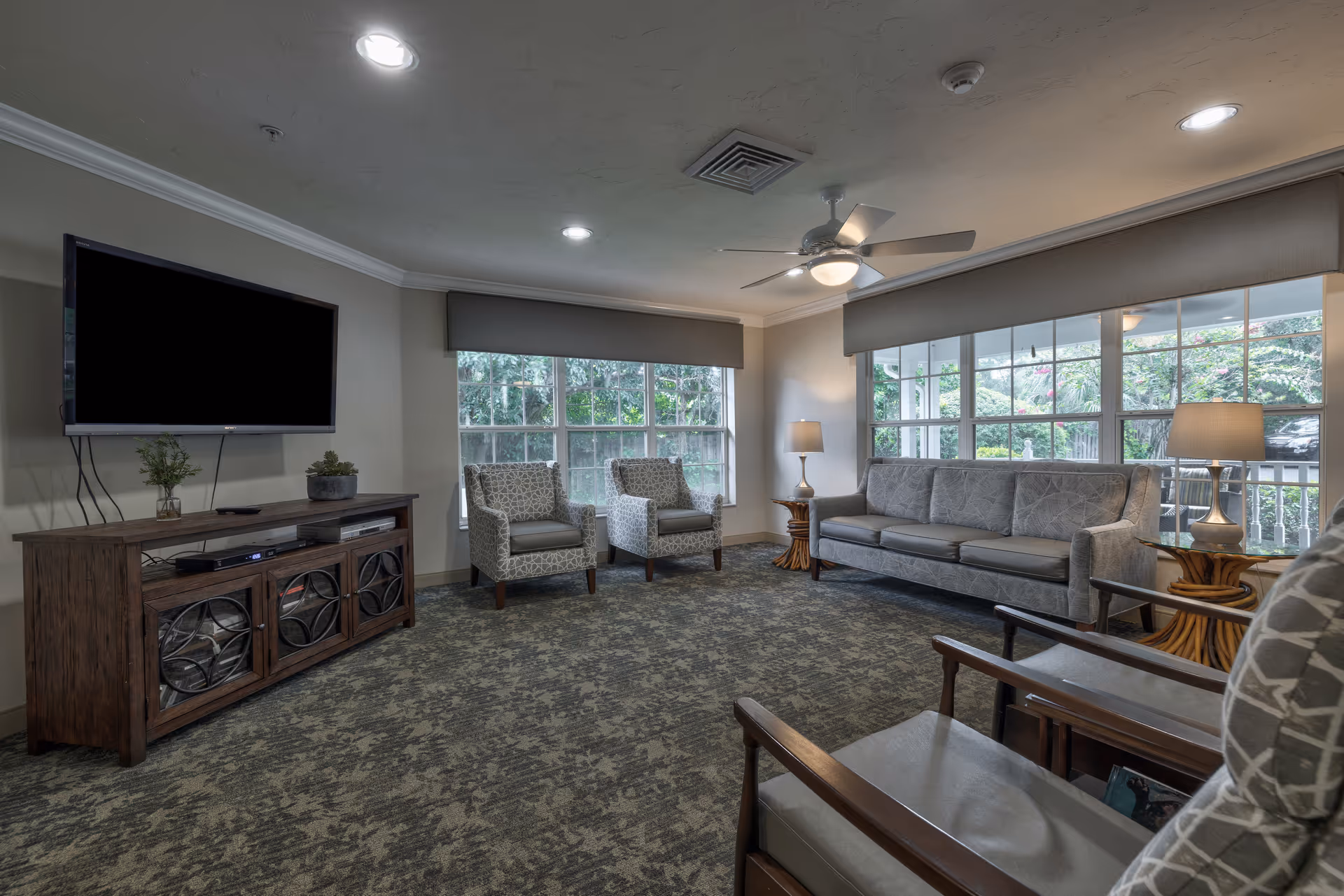 A cozy living room with a large window showing greenery outside. The room features a gray sofa, two patterned armchairs, a wooden TV stand with a flat-screen TV mounted above it, two side tables with lamps, and a ceiling fan with a light. The carpet has a subtle floral pattern, and the walls are painted a soft neutral color.