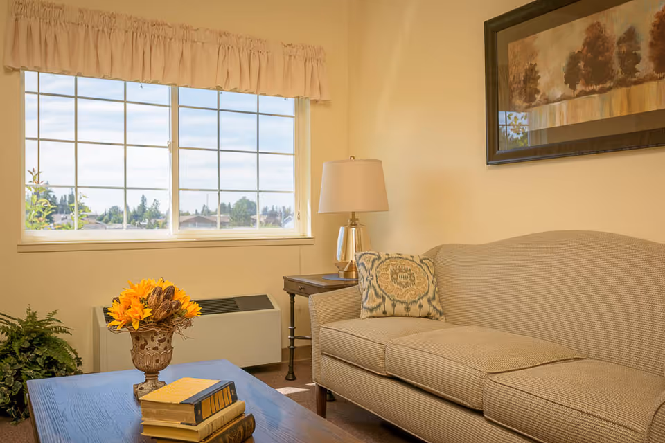 A cozy living room with a beige couch adorned with a decorative pillow, a wooden coffee table with a vase of sunflowers and stacked books, a side table with a lamp, a large window with a view of trees and houses outside, and a framed painting on the wall.