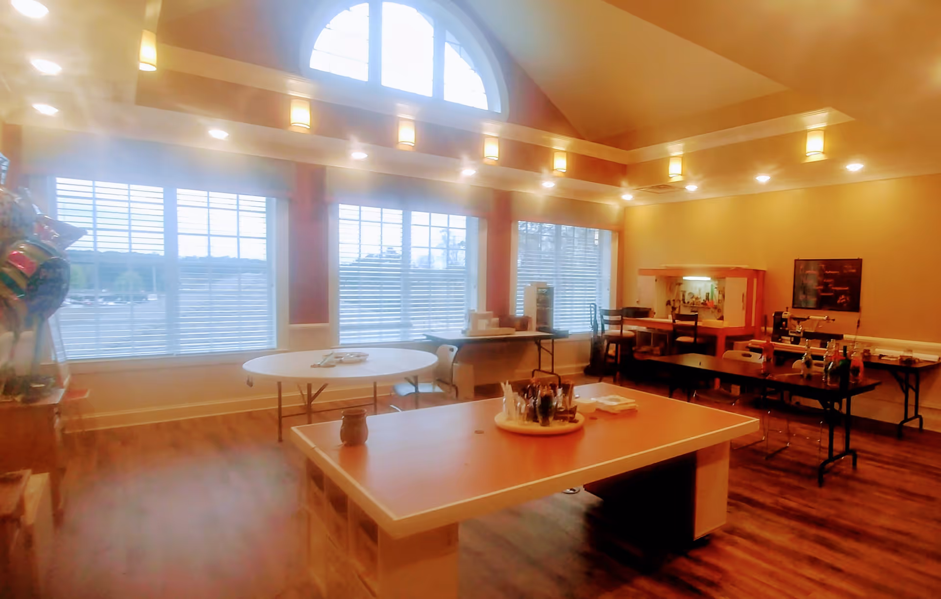 A spacious, well-lit activity or common room with large windows featuring white blinds. The room has a high ceiling with recessed lighting and wall sconces. There are several tables, including a large central table with art supplies and smaller tables around the room. The floor is wooden, and the walls are painted a warm beige color.