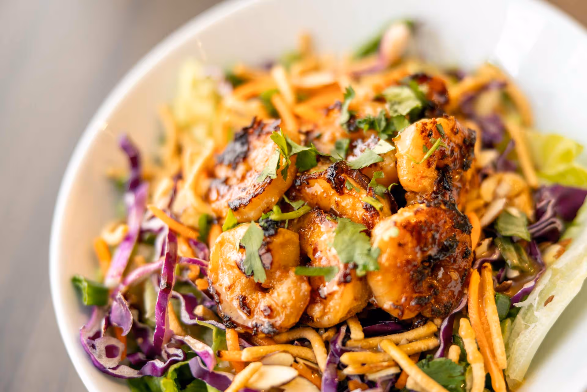 Close-up of a bowl of grilled shrimp served on a bed of shredded purple cabbage, lettuce, carrots, and garnished with chopped cilantro and crunchy noodles.
