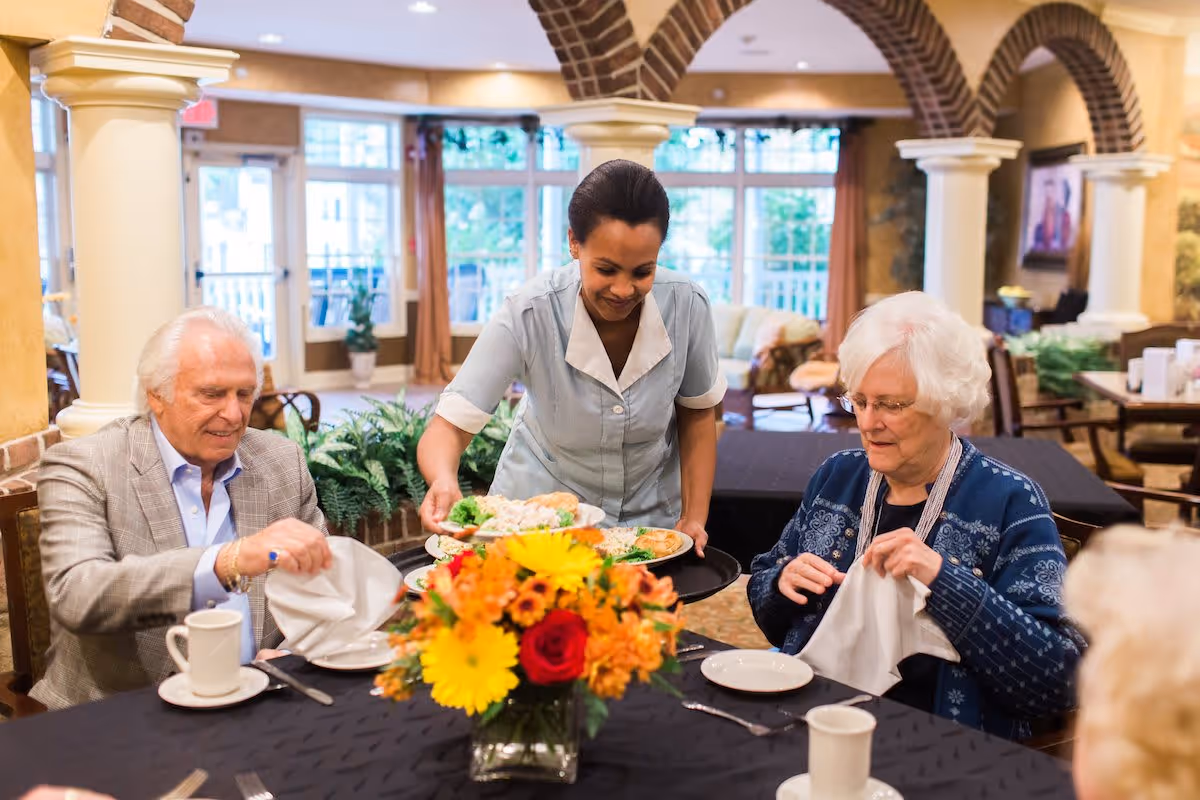 A dining room in a senior living facility where a caregiver is serving food to elderly residents seated at a table with a colorful flower centerpiece. The room has large windows, brick arches, and a warm, inviting atmosphere.