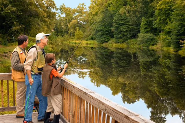 Three people fishing from a wooden deck overlooking a calm pond surrounded by green trees. An older man, a middle-aged man, and a young boy are engaged in fishing, with the boy holding a fishing rod.
